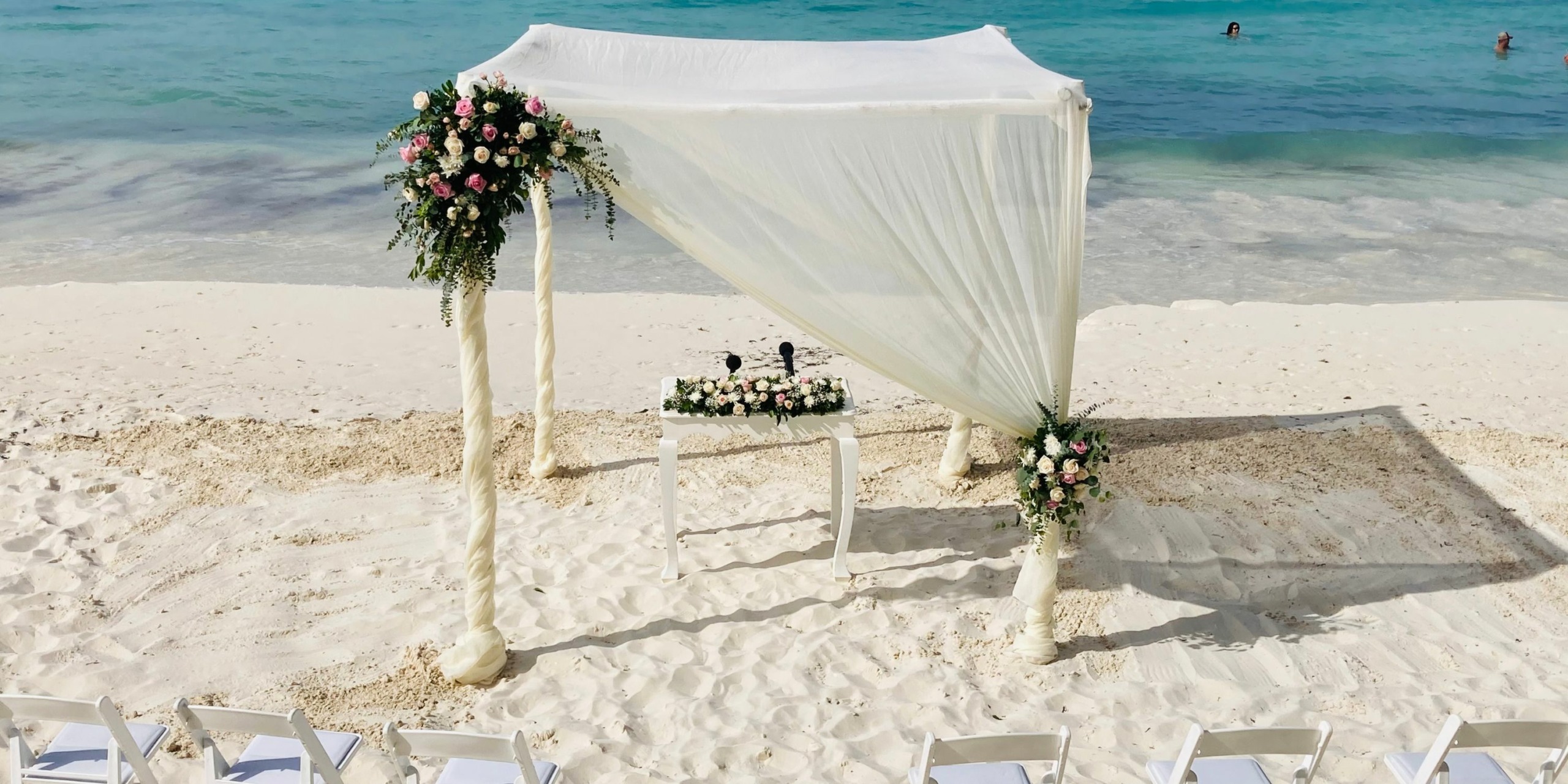 A destination beach wedding setup with a white canopy decorated with flowers, accompanied by a matching floral table and white chairs on sandy shore.