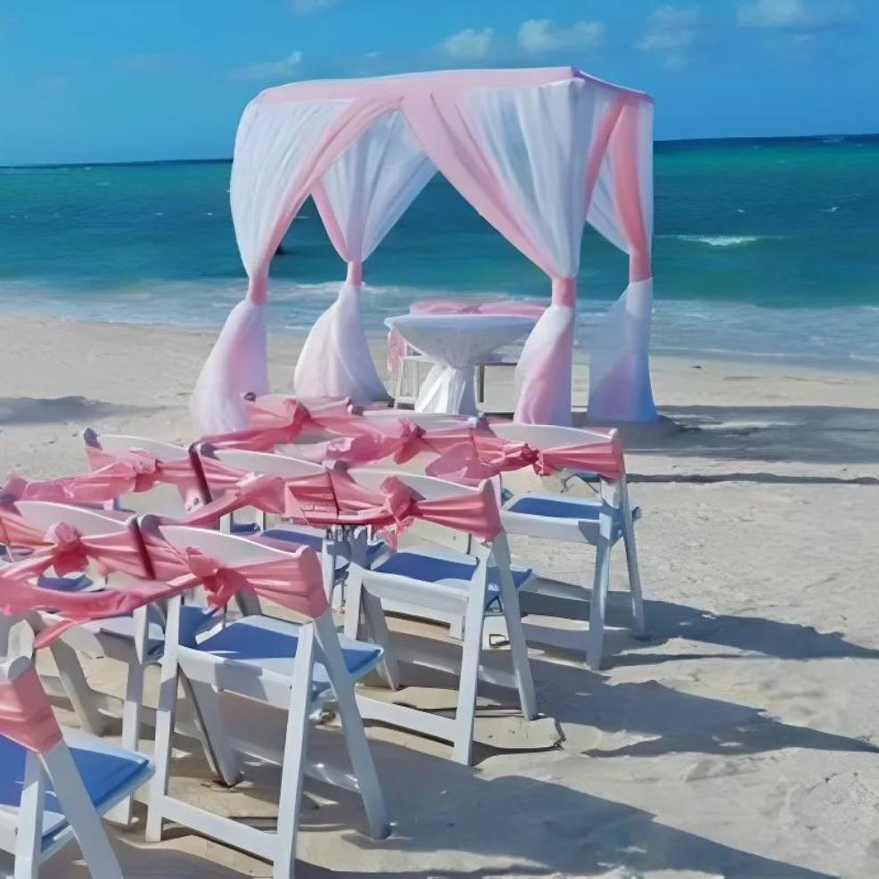 Destination beach wedding setup with white chairs adorned with pink ribbons and a canopy draped in pink and white, facing the turquoise sea.