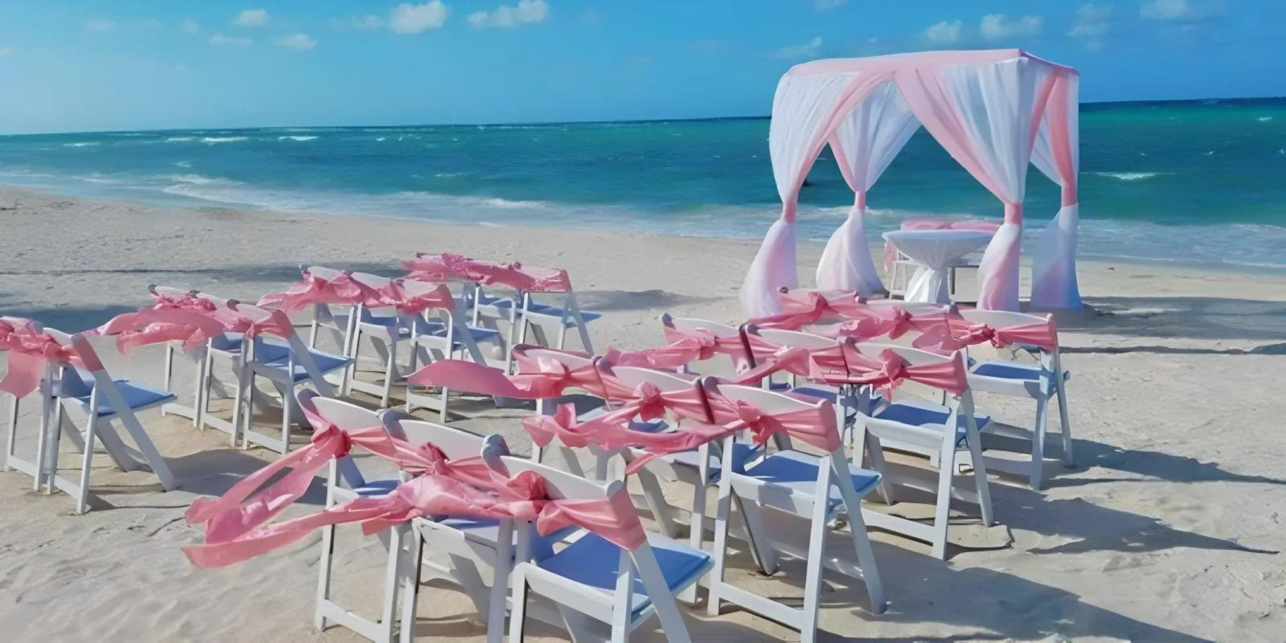 White chairs with pink ribbons set up for a destination wedding ceremony under a white canopy by the sea.