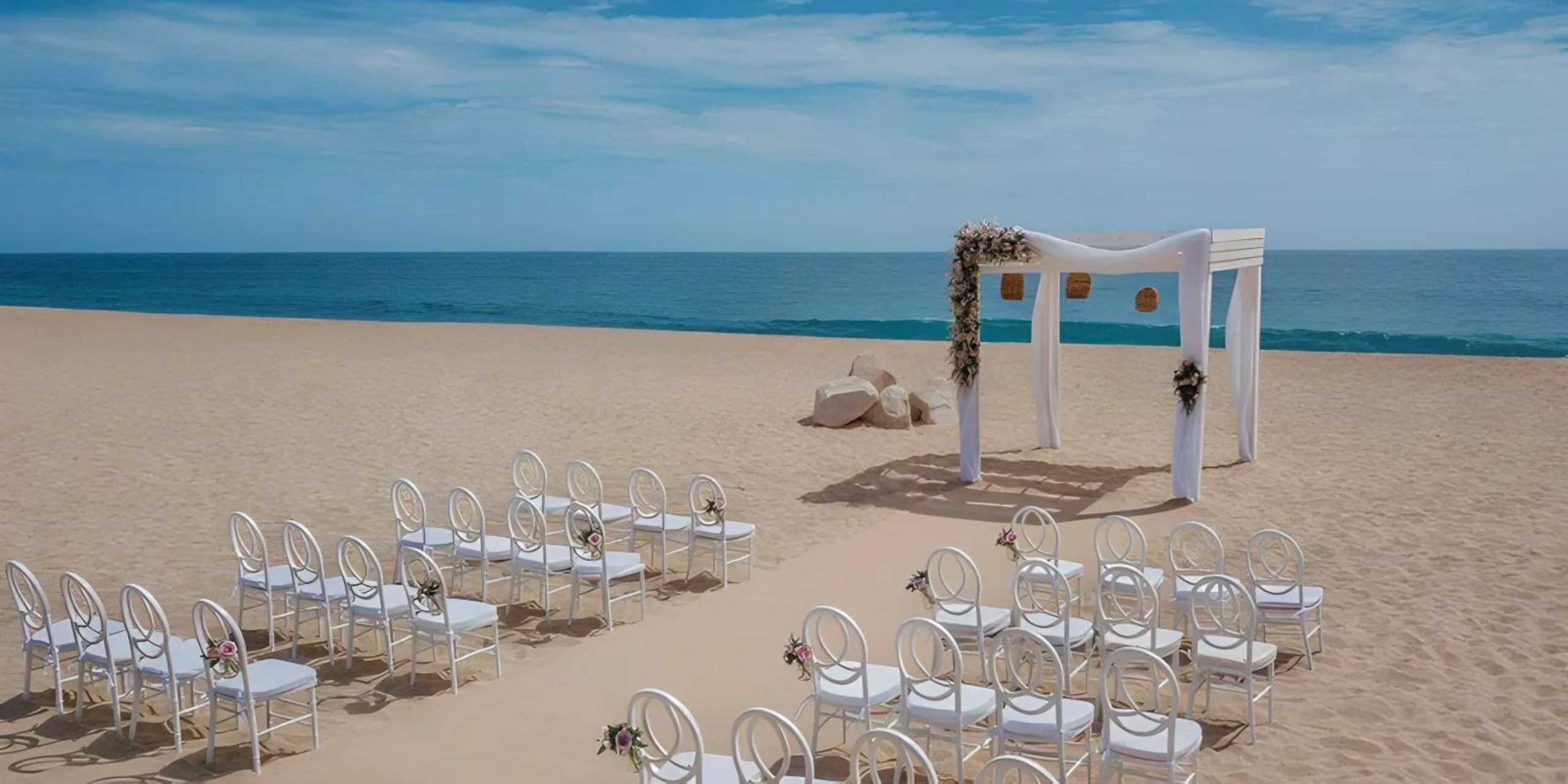 Destination beach wedding setup with white chairs arranged in rows facing an altar decorated with flowers, under a clear blue sky.