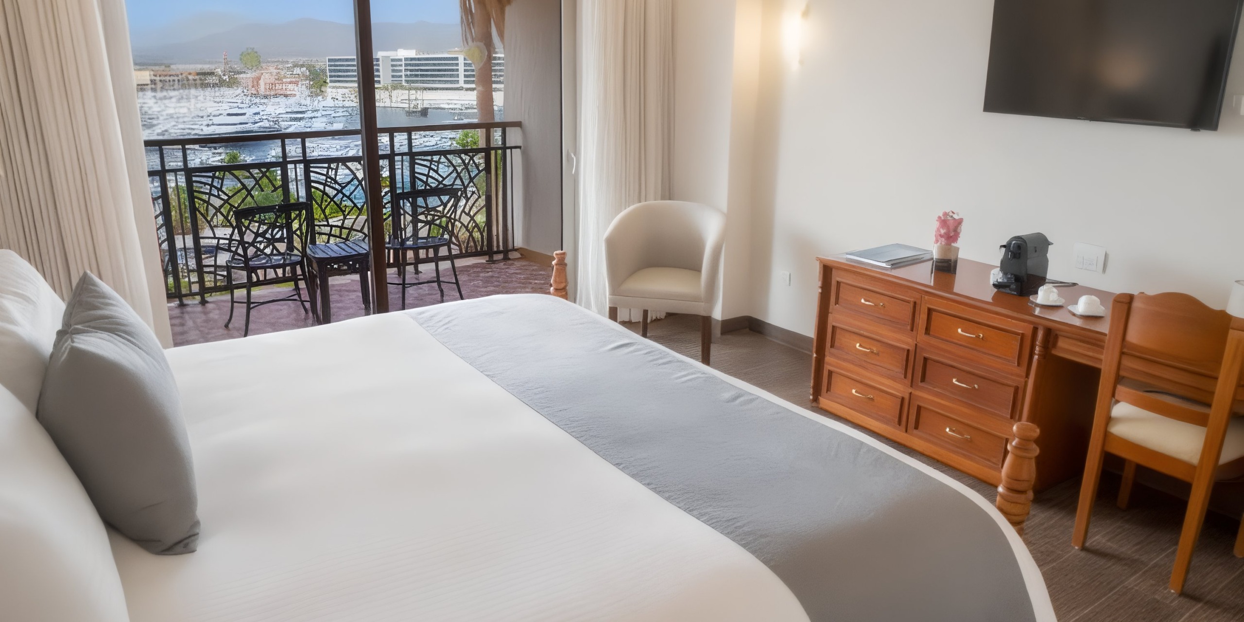 Enjoy a room at Sandos Finisterra Los Cabos featuring a large bed, a chair, a dresser with a TV and coffee maker, and a balcony with stunning views of the marina.