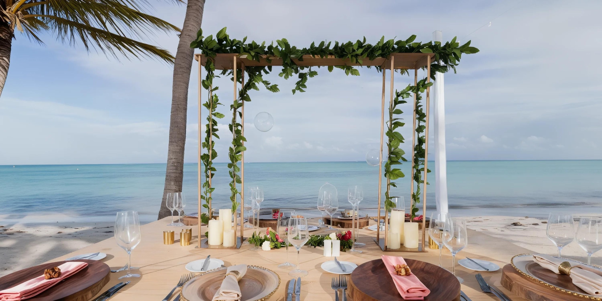 A Secrets Cap Cana wedding table setup on the beachfront, adorned with wooden furniture, lush greenery, elegant candles, and stylish glasses. The ocean serves as a stunning backdrop under a partly cloudy sky.