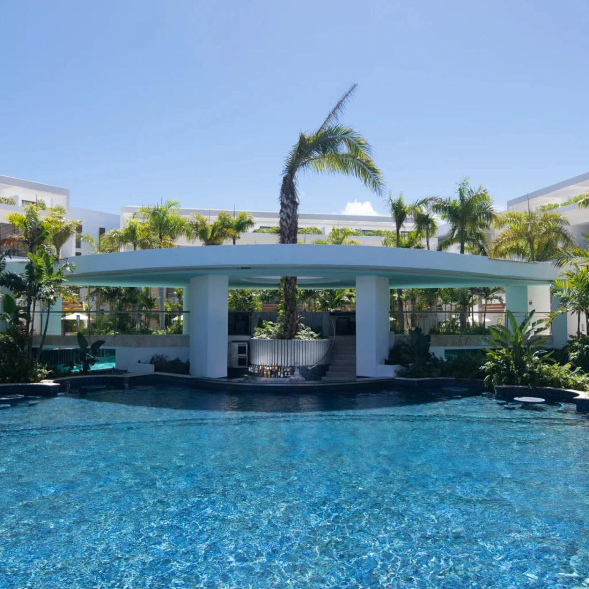 Sentence with product name: A luxurious pool with a swim-up bar shaded by a modern white pavilion at Grand Palladium Costa Mujeres, surrounded by lush vegetation and palm trees under a clear blue sky, perfect for a destination wedding.