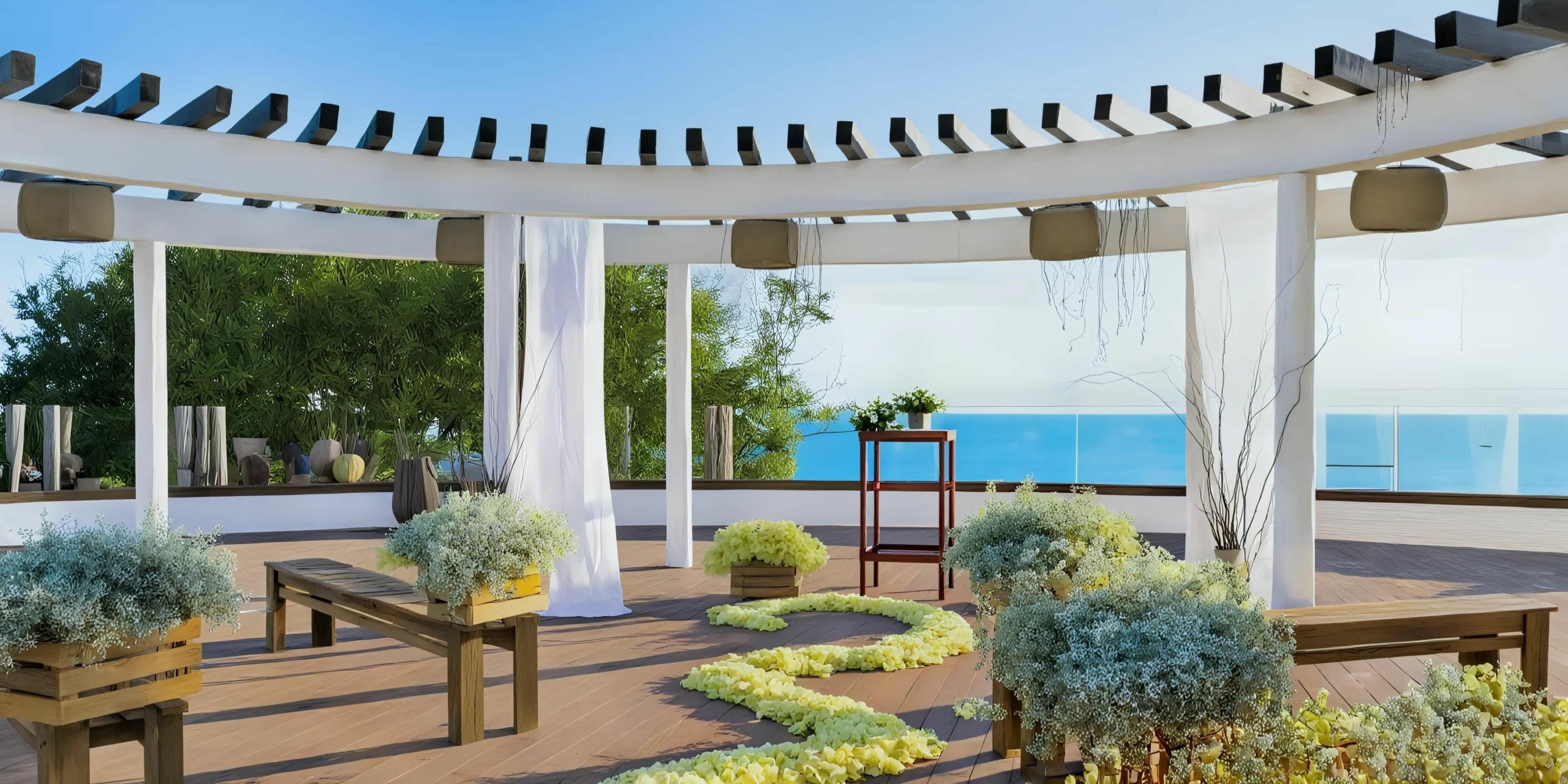 An outdoor wedding setup at the Grand Palladium Vallarta, featuring wooden benches, floral arrangements, and a pergola with white drapery, overlooking a clear blue ocean.