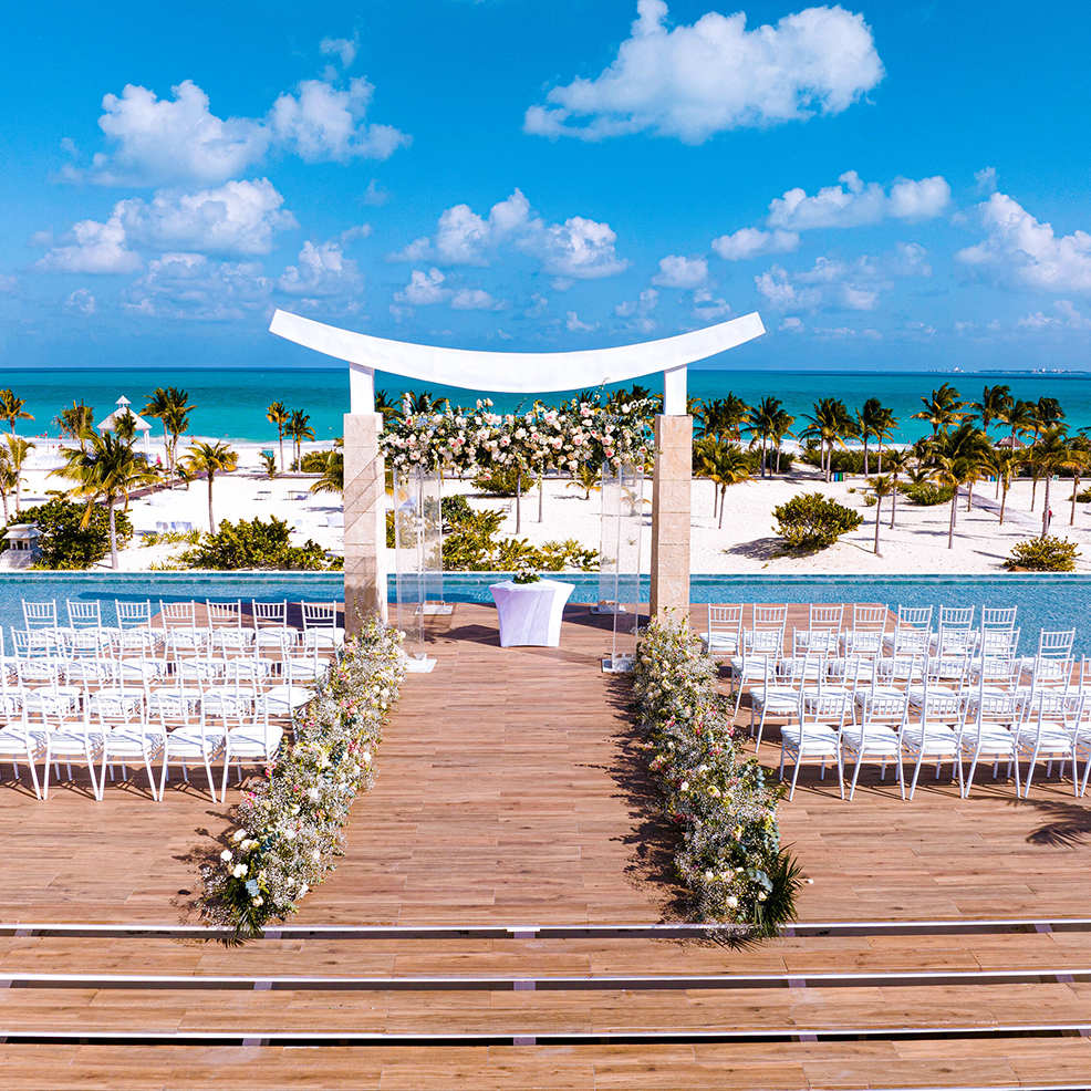 Wooden outdoor wedding aisle with flower decorations, facing an altar and the ocean. White chairs arranged on both sides, palm trees and a blue sky in the background.