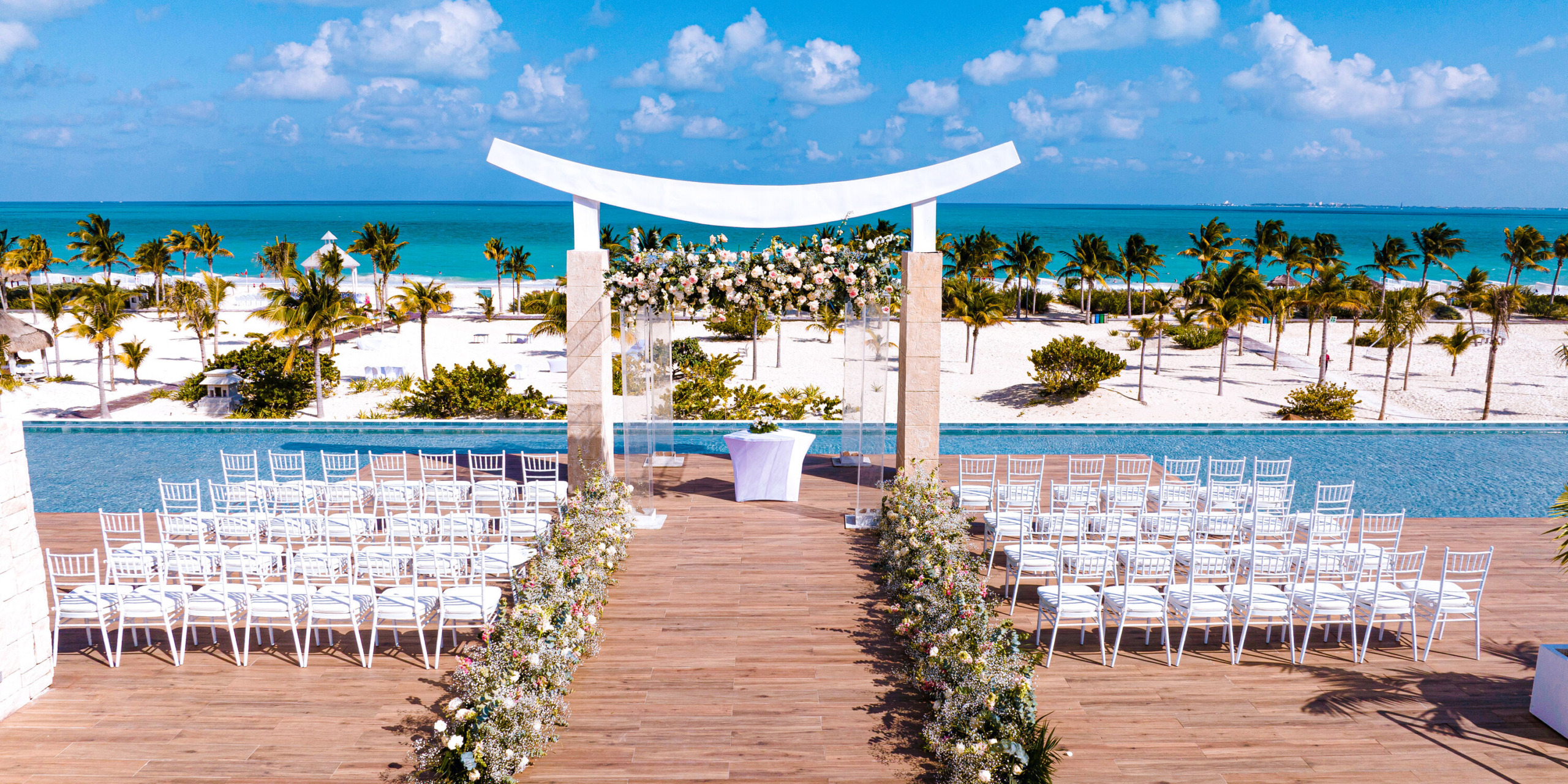 The outdoor wedding at Majestic Elegance Costa Mujeres features an ocean-facing setup with rows of white chairs, a beautifully decorated archway, and a picturesque backdrop of palm trees and blue sky.