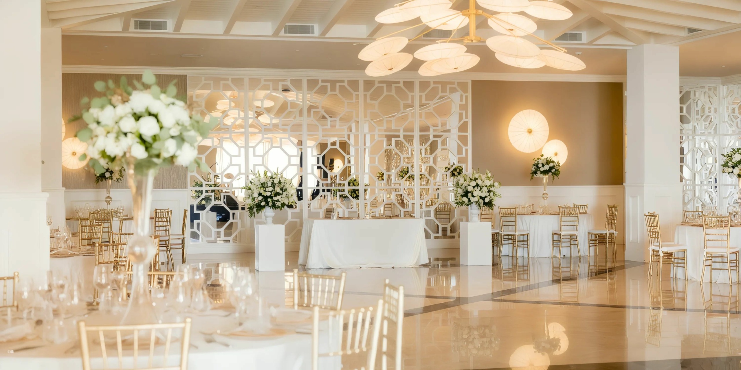 Majestic Elegance Costa Mujeres features a refined banquet hall adorned with white floral arrangements, gold chairs, and a reflective floor. The room's sophistication is further accentuated by geometric wall designs and contemporary lighting fixtures.