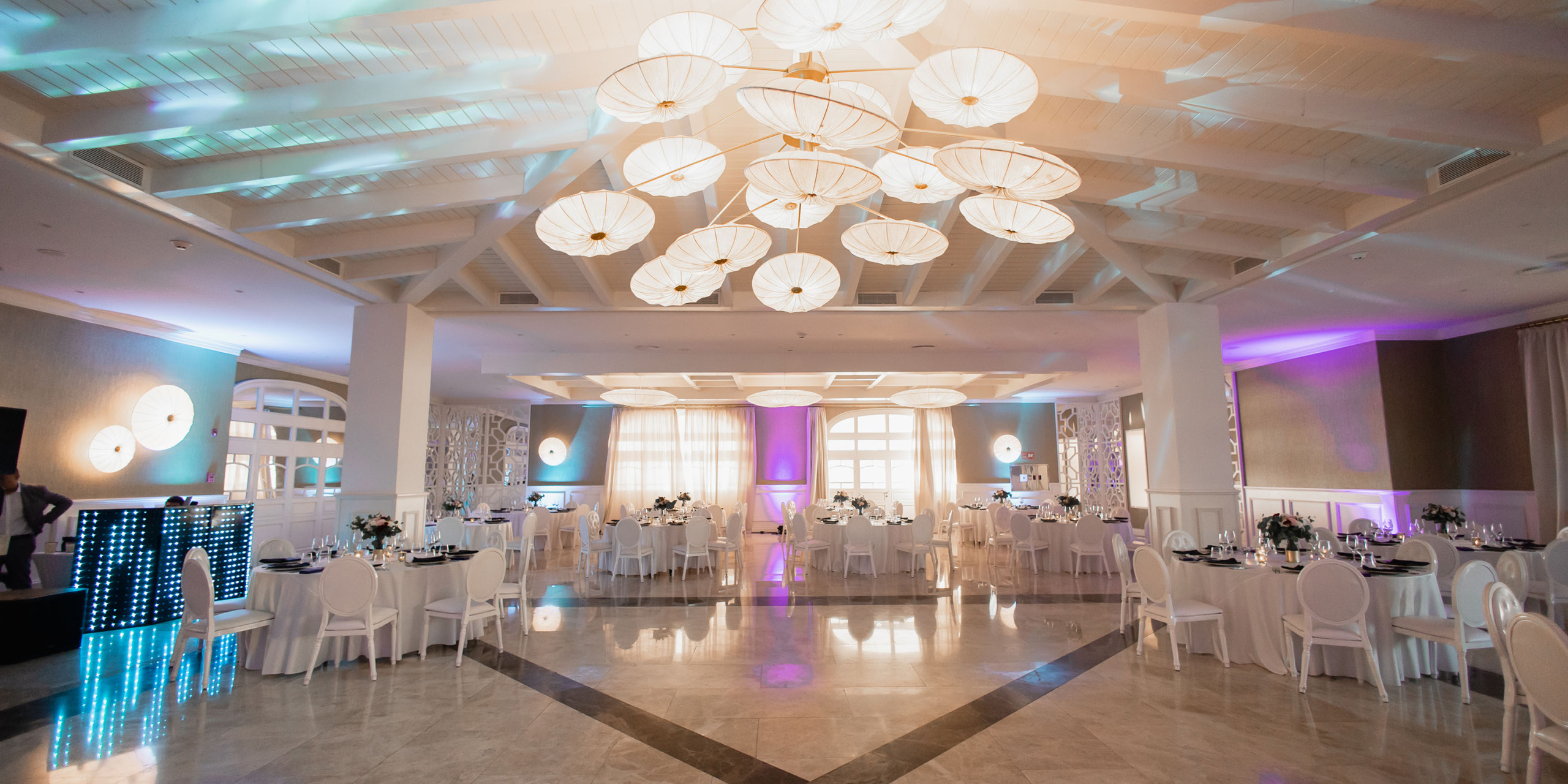 Elegant Majestic Elegance Costa Mujeres wedding reception hall decorated with white tables, chairs, and hanging umbrellas from the ceiling, illuminated by soft pink lighting.