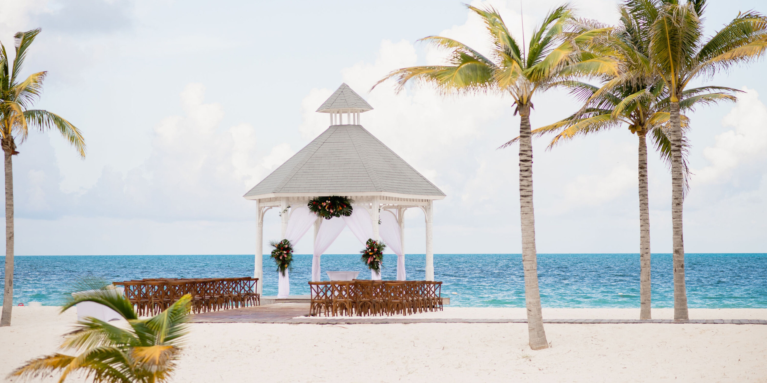The Majestic Elegance Costa Mujeres offers a beachside gazebo with floral decorations, wooden seating, and palm trees, all overlooking the ocean under a cloudy sky.