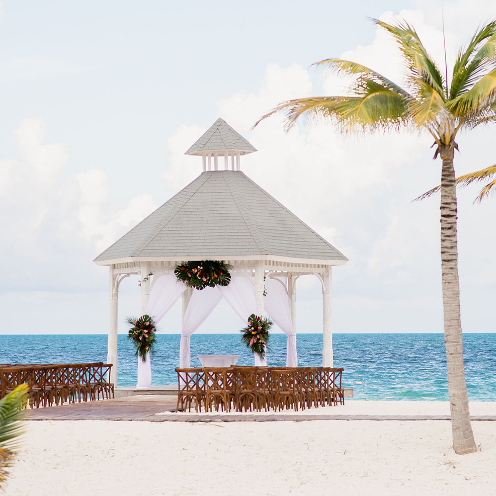 A white gazebo with floral decorations overlooks a sandy beach and blue ocean. Rows of wooden chairs are set up facing the gazebo. Palm trees are visible on the right.