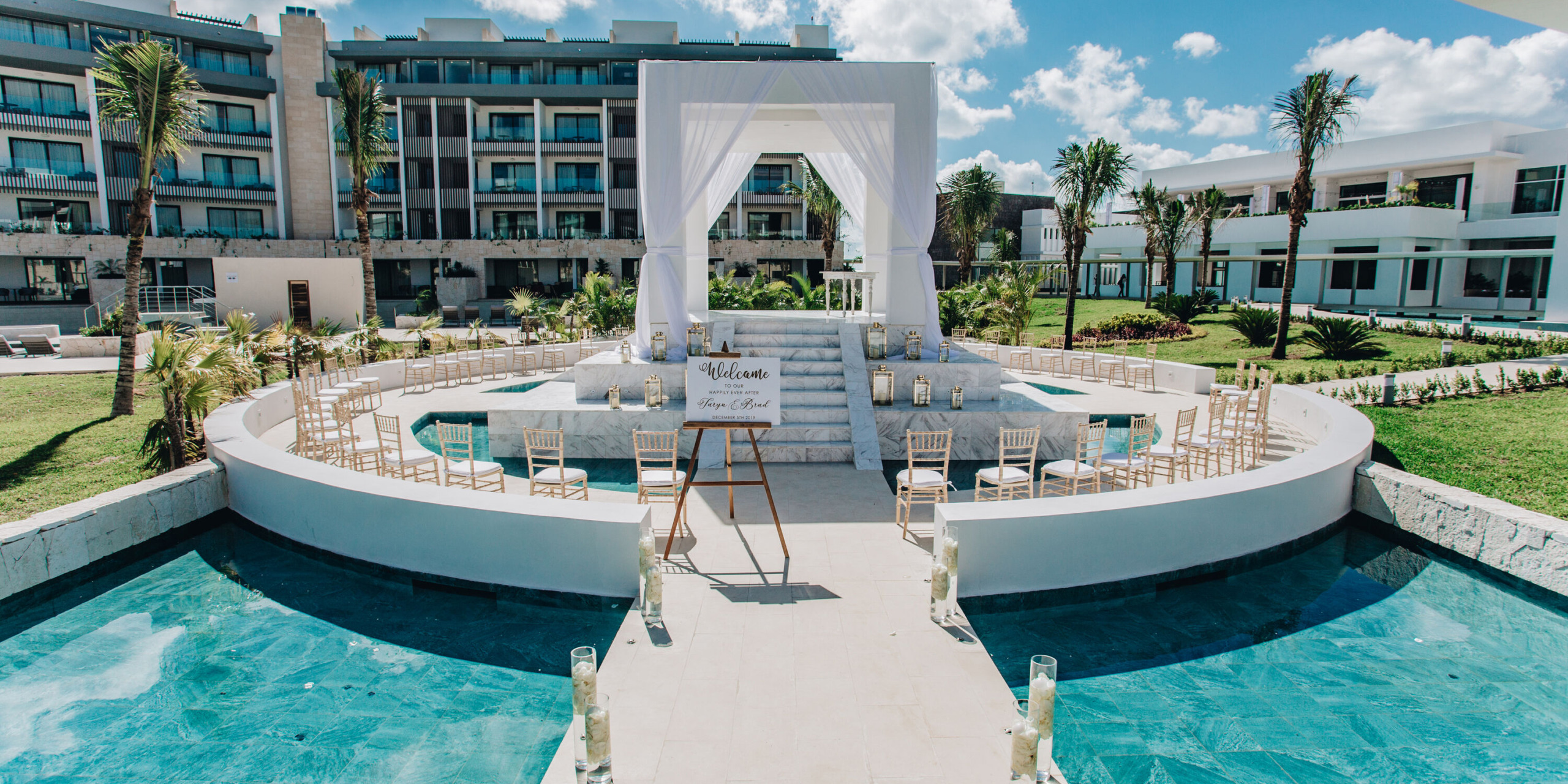 The Majestic Elegance Costa Mujeres features an outdoor wedding venue with a central altar, surrounded by semi-circular seating in landscaped grounds with a pool, all set beneath a partly cloudy sky.