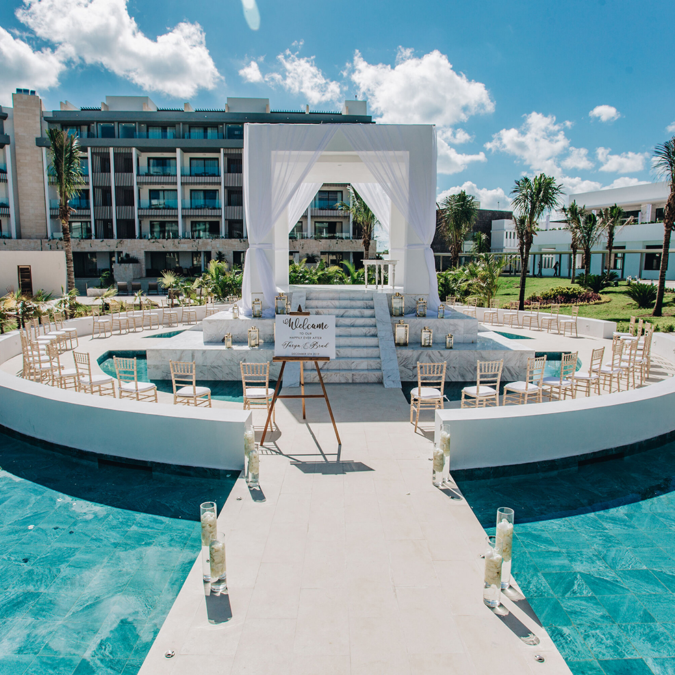 Outdoor wedding setup with a white arch, chairs arranged around a circular platform, and a sign reading "Welcome." A modern building and palm trees are in the background.