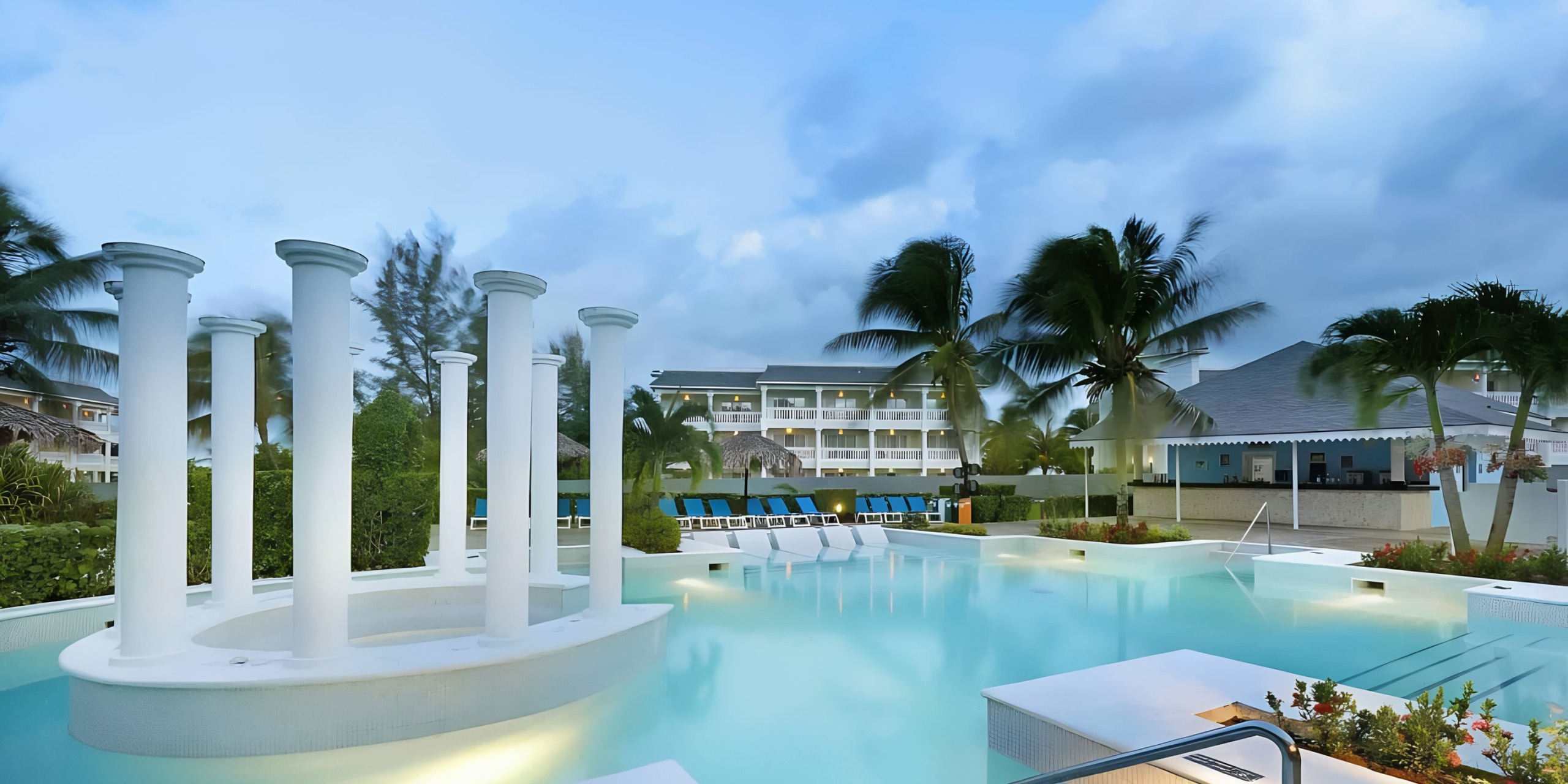 A serene Grand Palladium Lady Hamilton resort pool at dusk with classical columns and tropical palm trees.