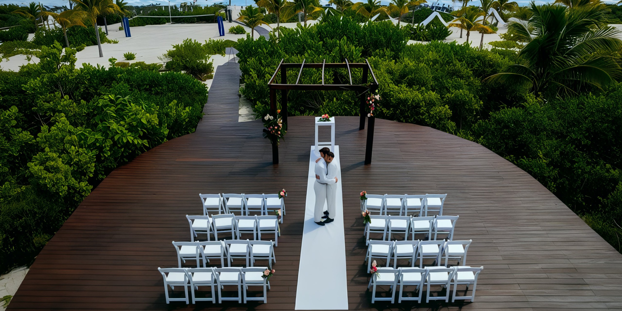 A groom in a white suit stands under a pergola on a wooden deck at the Grand Palladium Costa Mujeres, with rows of white chairs arranged for an outdoor wedding ceremony amidst lush greenery.