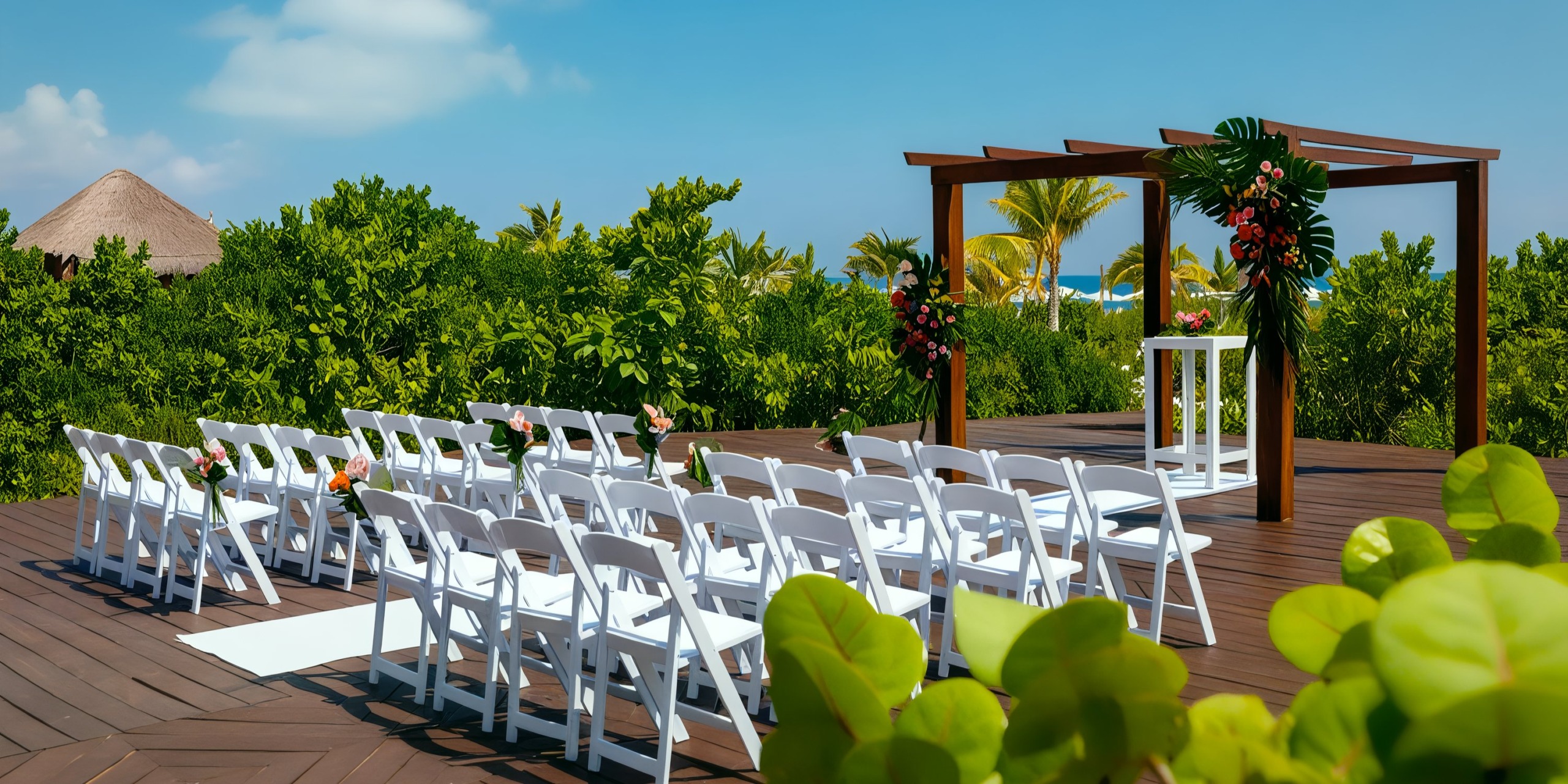 Experience the Grand Palladium Costa Mujeres for your outdoor wedding, featuring rows of white chairs facing a wooden arch adorned with flowers. The venue is surrounded by lush greenery, with a charming thatched roof providing an enchanting backdrop.
