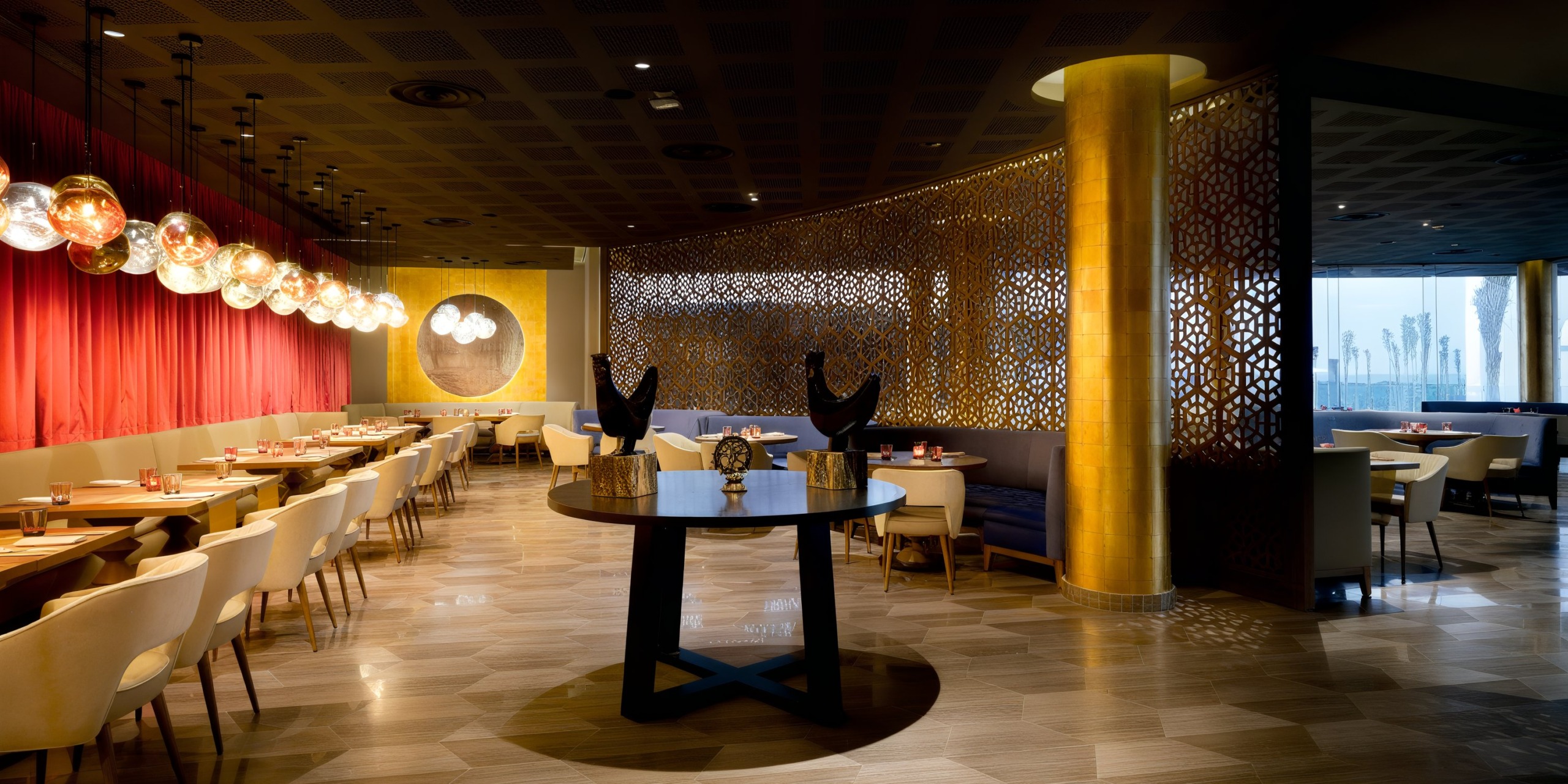 The Grand Palladium Costa Mujeres offers a sophisticated and stylish restaurant interior, boasting red curtains, circular hanging lights, wooden tables, cream chairs, and gold decorative accents with a geometric latticework partition.