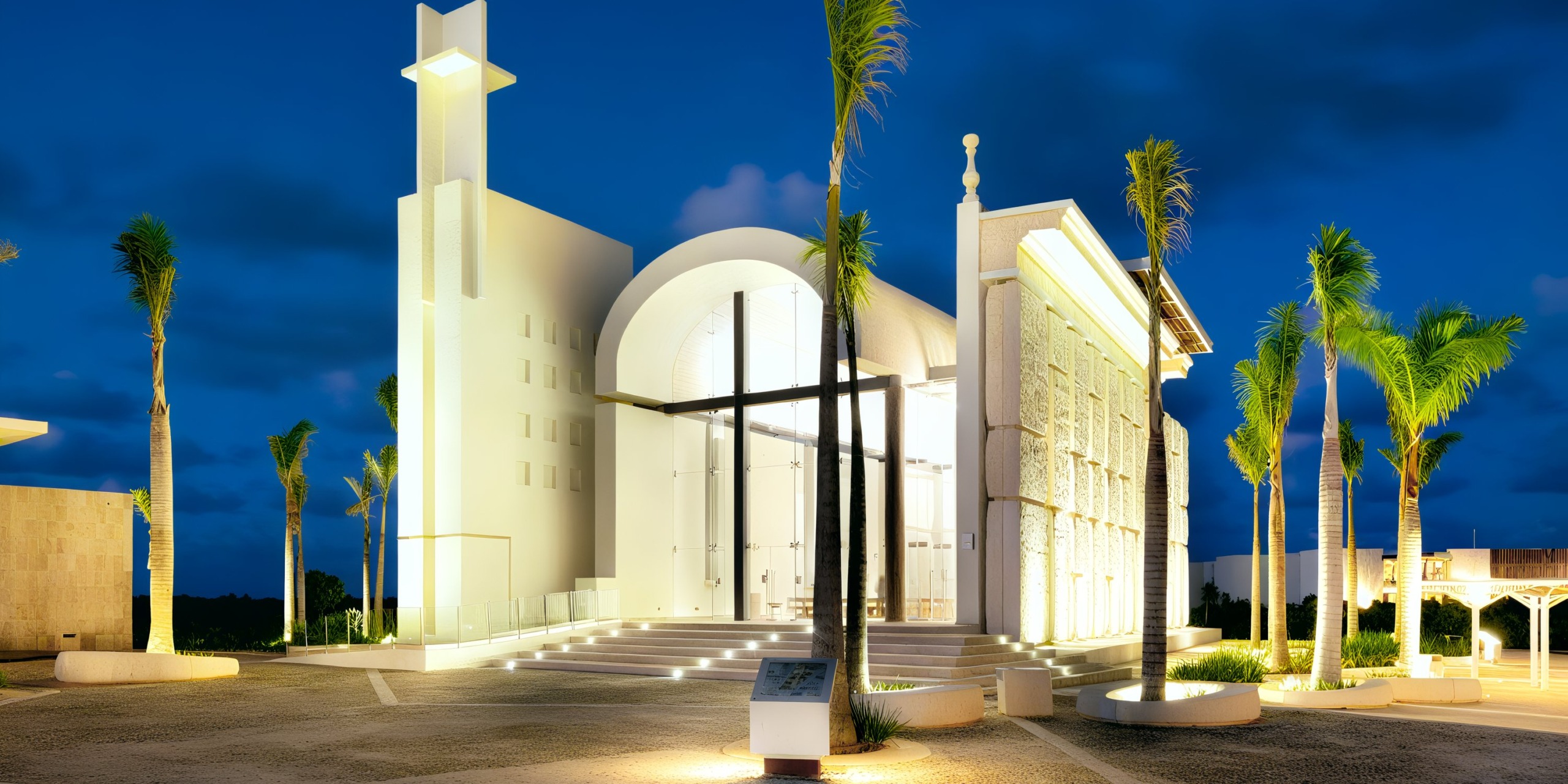 The Grand Palladium Costa Mujeres features a modern architectural design with a prominent illuminated cross at its entrance, beautifully enhanced by surrounding palm trees.