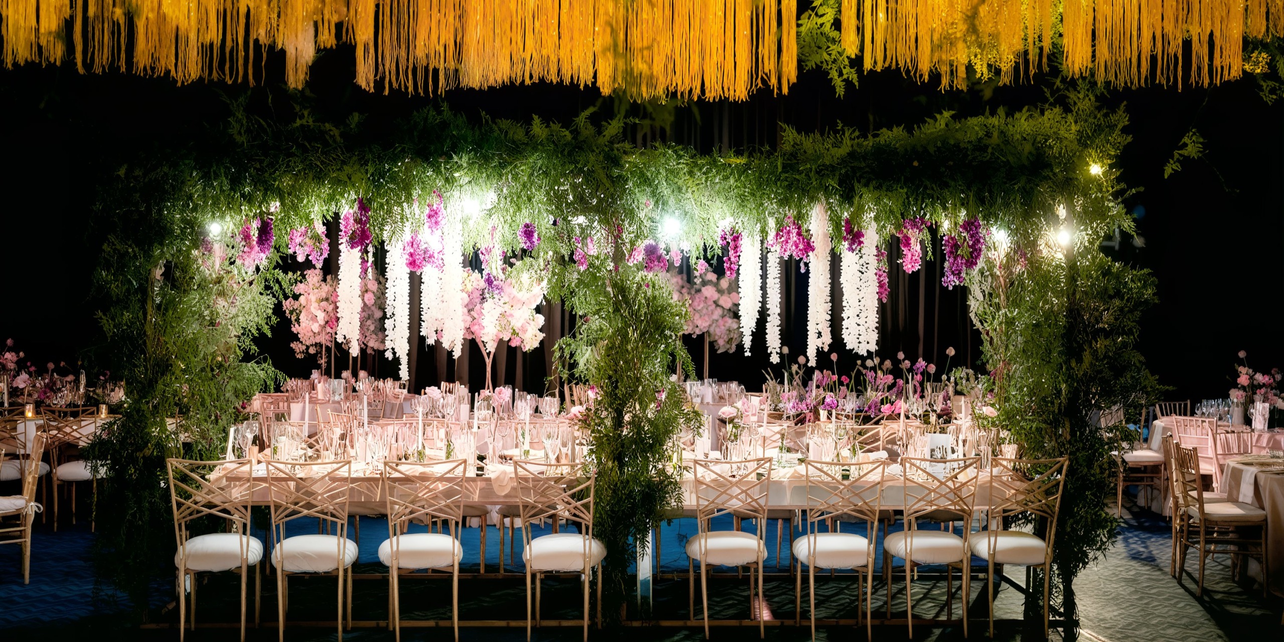 Immerse yourself in an elegant outdoor event setup at the Grand Palladium Costa Mujeres, featuring a long table adorned with pristine white chairs, vibrant hanging yellow and purple flowers, and lush greenery.