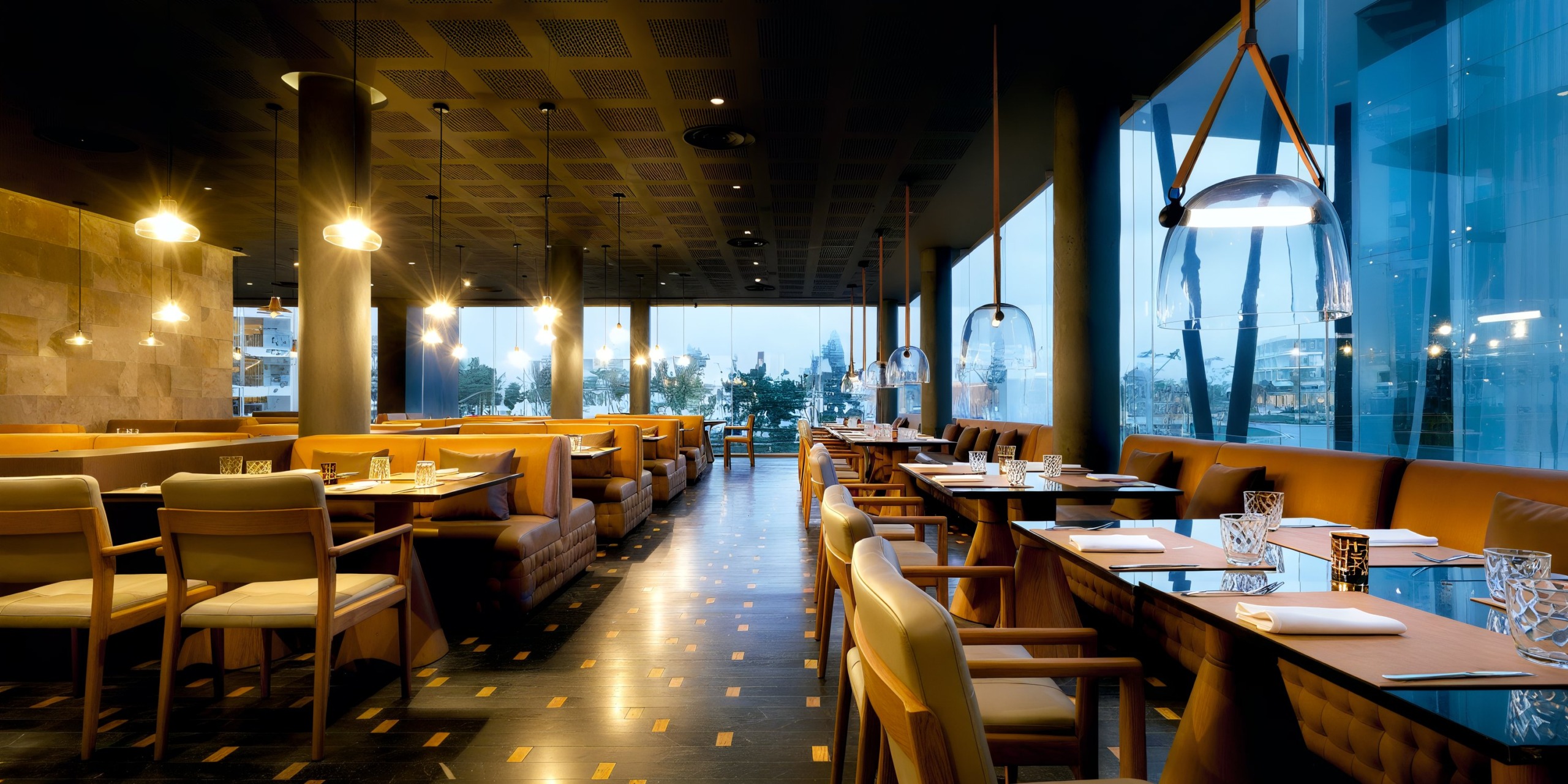 A modern restaurant interior at Grand Palladium Costa Mujeres features wooden tables and chairs, leather booths, and large windows offering a city view. The warm lighting adds to the cozy atmosphere.