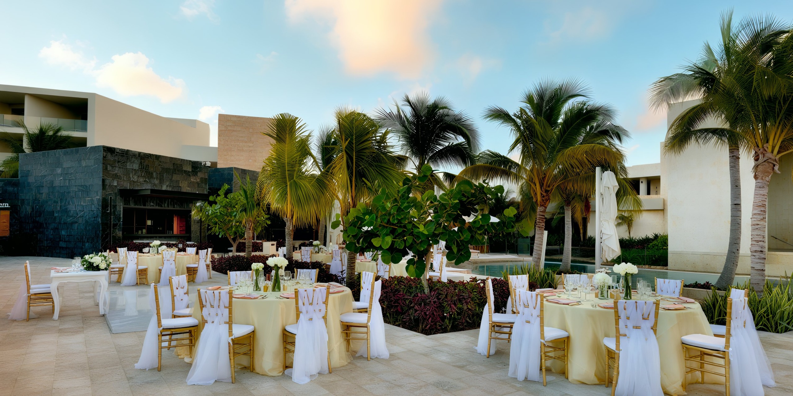 The Grand Palladium Costa Mujeres outdoor wedding reception features round tables and white chairs elegantly adorned with white and gold tableware, set amidst a backdrop of palm trees and modern buildings under a clear blue sky.