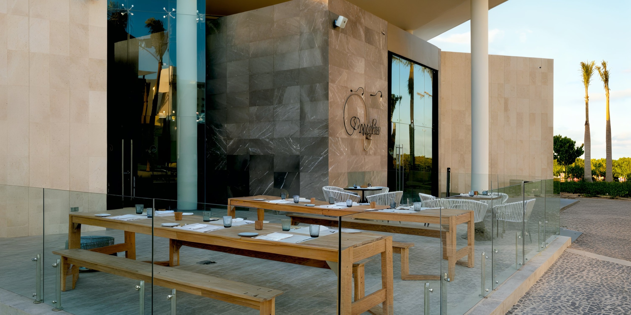 An outdoor dining area featuring wooden tables and benches prepared for service, enclosed by glass fencing, is situated in front of a contemporary building with expansive windows and a sign that reads "Grand Palladium Costa Mujeres.