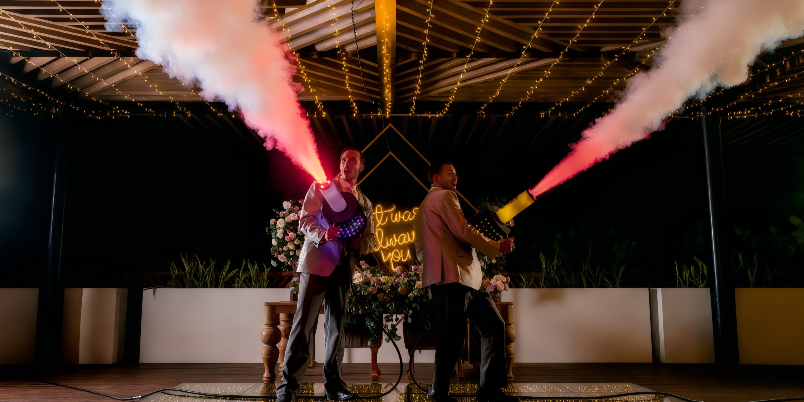 Two people are holding smoke machines emitting red smoke at the Grand Palladium Costa Mujeres outdoor stage, which is adorned with string lights and floral decorations.