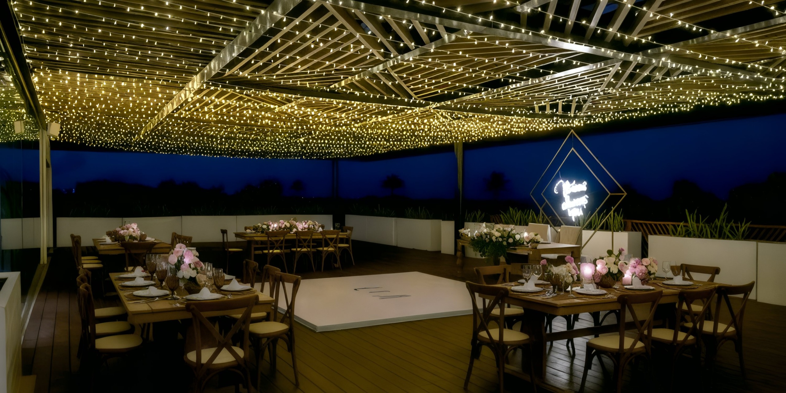 The outdoor event space at Grand Palladium Costa Mujeres features wooden tables, cushioned chairs, flower arrangements, and a central dance floor. Overhead, string lights and a neon sign illuminate the night, creating an inviting ambiance.