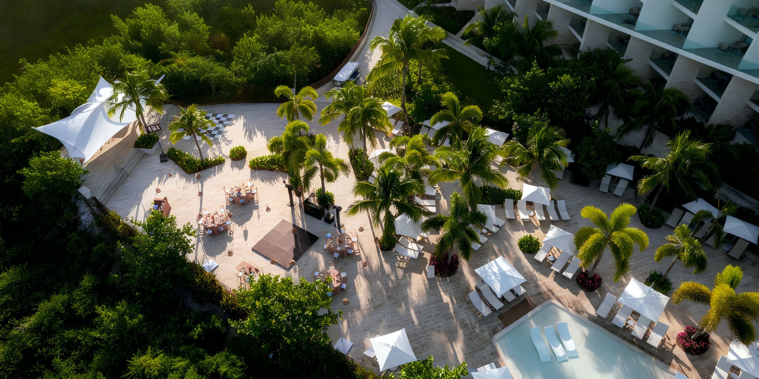 Aerial view of the sunlit Grand Palladium Costa Mujeres resort area featuring palm trees, lounge chairs, tables, and umbrellas surrounding a pool.