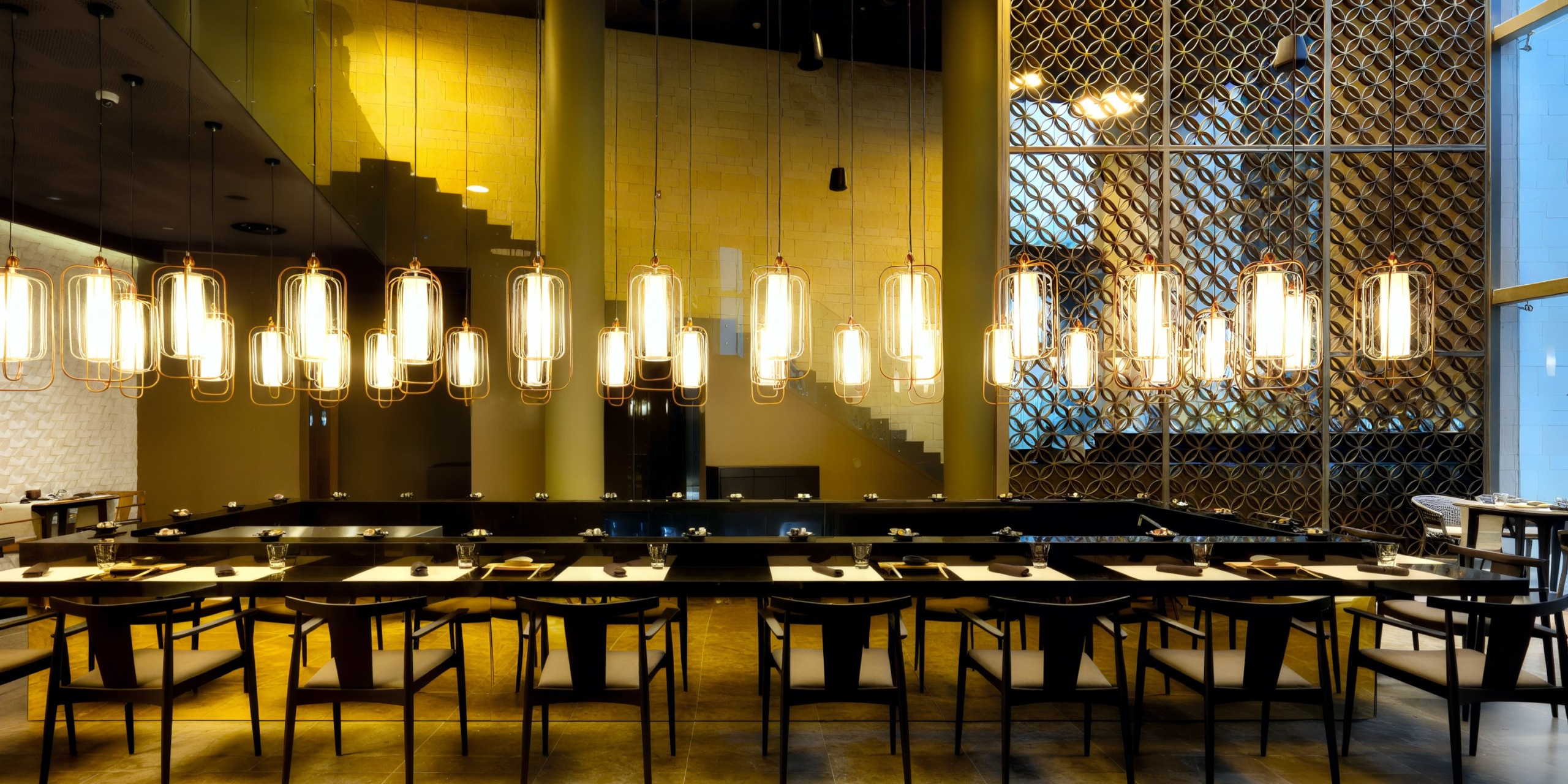 The Grand Palladium Costa Mujeres showcases a contemporary restaurant interior complete with a long table surrounded by chairs and illuminated by hanging pendant lights, all set against a backdrop of expansive windows and decorative wall panels.