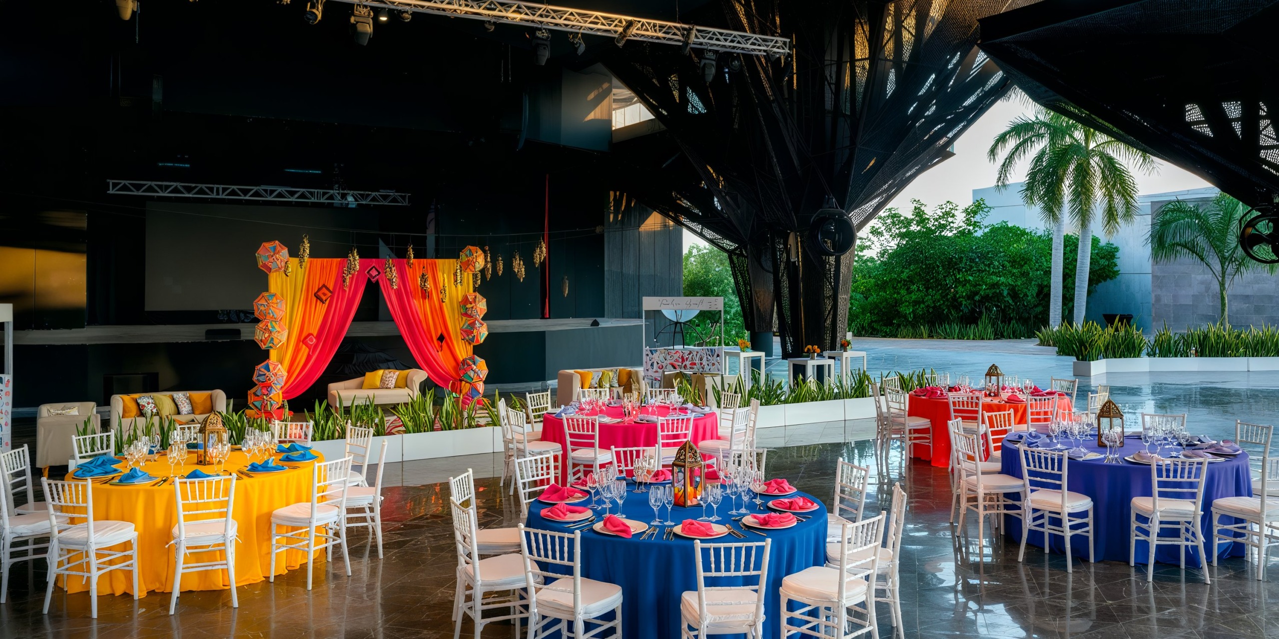 Host your outdoor event with round tables adorned in vibrant tablecloths and matching chairs at the Grand Palladium Costa Mujeres. A decorative stage featuring drapes and hanging ornaments serves as a captivating backdrop, complemented by the view of palm trees outside.