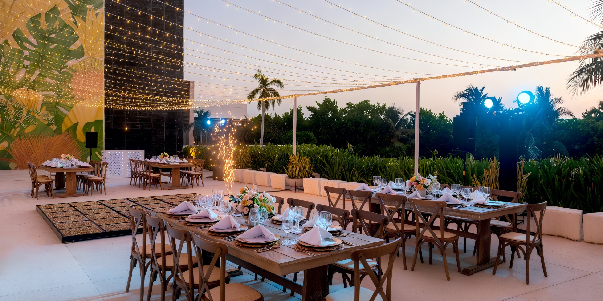 Experience a unique outdoor wedding reception at Grand Palladium Costa Mujeres, featuring elegant wooden tables and chairs adorned with white tablecloths and beautiful floral centerpieces. Delicate string lights sparkle overhead, while palm trees and lush greenery provide a stunning backdrop.