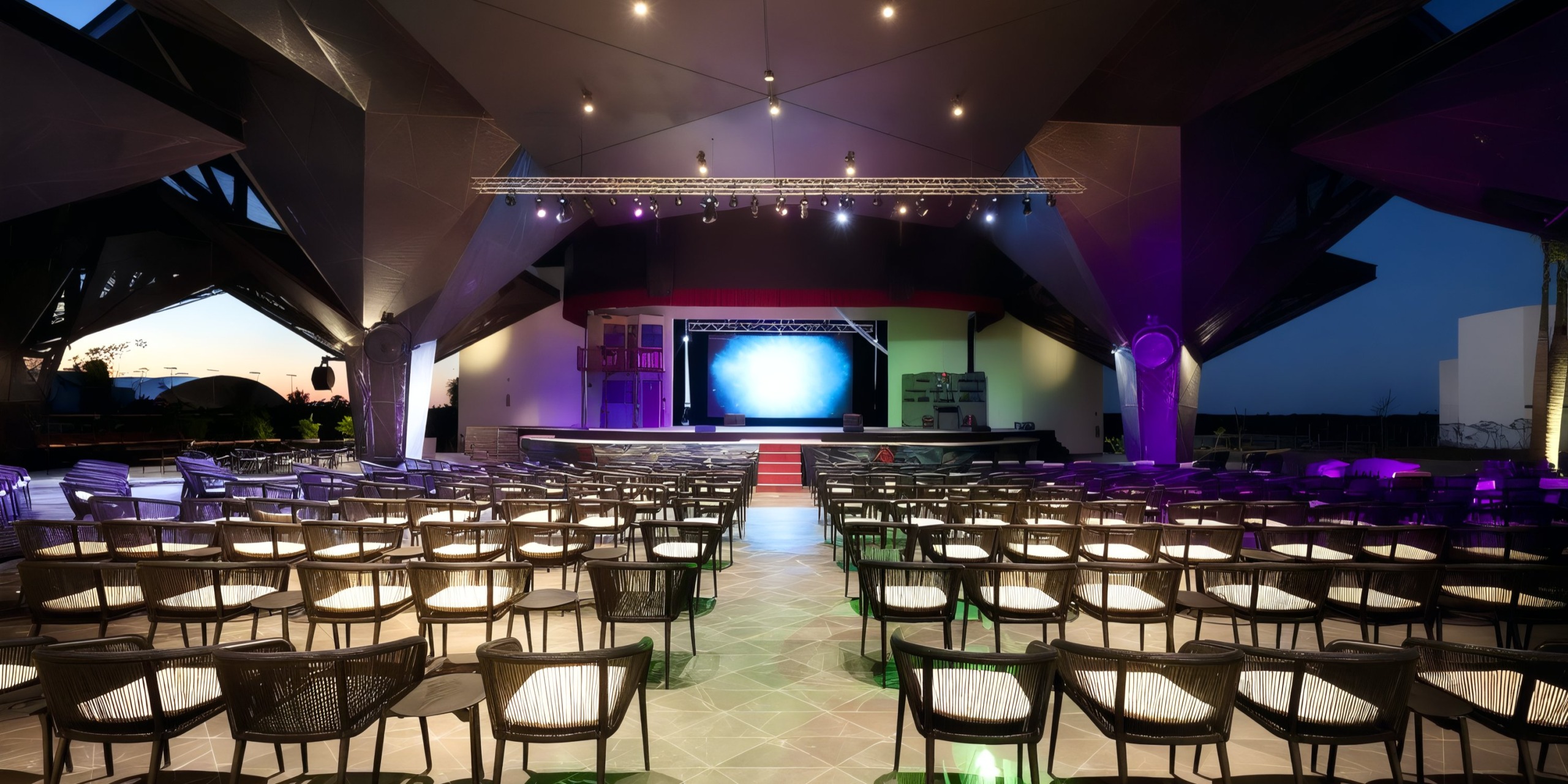 The Grand Palladium Costa Mujeres features an empty auditorium with rows of chairs facing a stage illuminated in shades of blue and purple, complemented by a ceiling design showcasing angular, modern architecture.