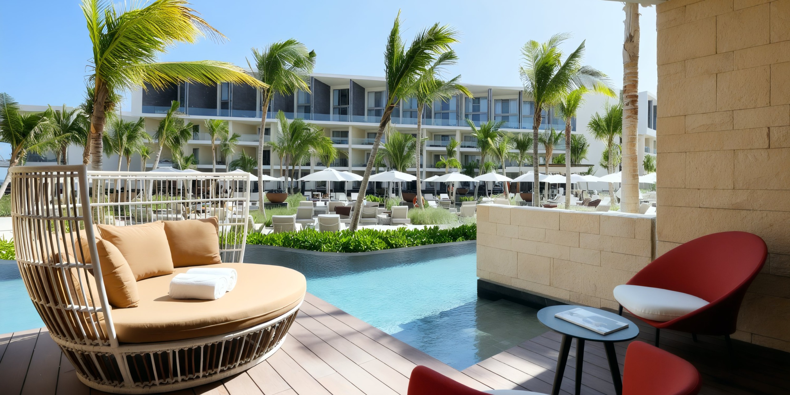 The scene features a poolside view at the Grand Palladium Costa Mujeres, complete with lounge chairs, a daybed, and red chairs beside a small pool, all surrounded by palm trees and modern hotel architecture in the background.
