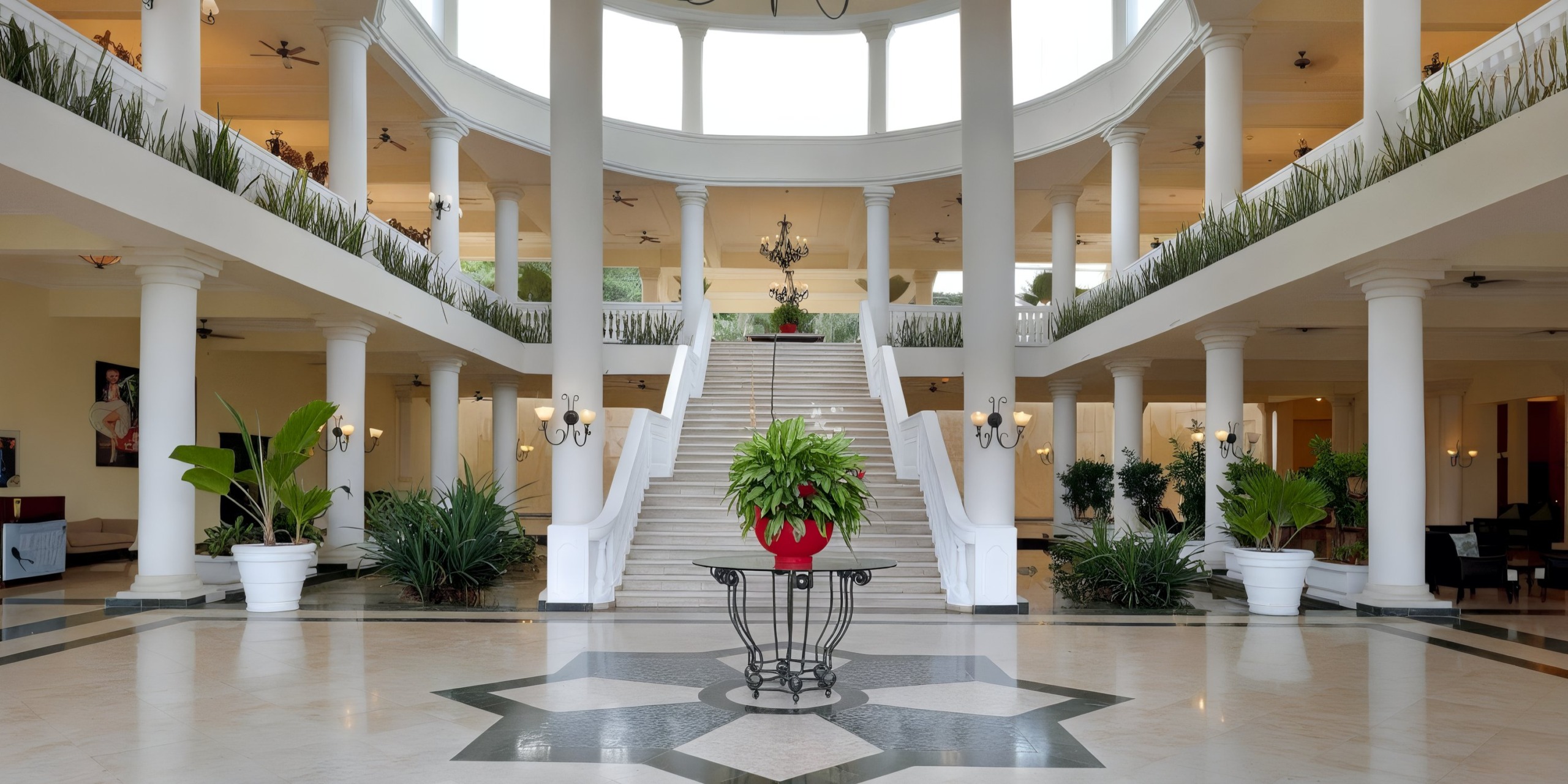 The Grand Palladium Lady Hamilton exudes sophistication, showcasing a magnificent lobby with an impressive staircase, white columns, vibrant greenery, and a central table highlighted by a red planter. Its lofty ceiling and plentiful natural light create the perfect backdrop for a breathtaking wedding ceremony.