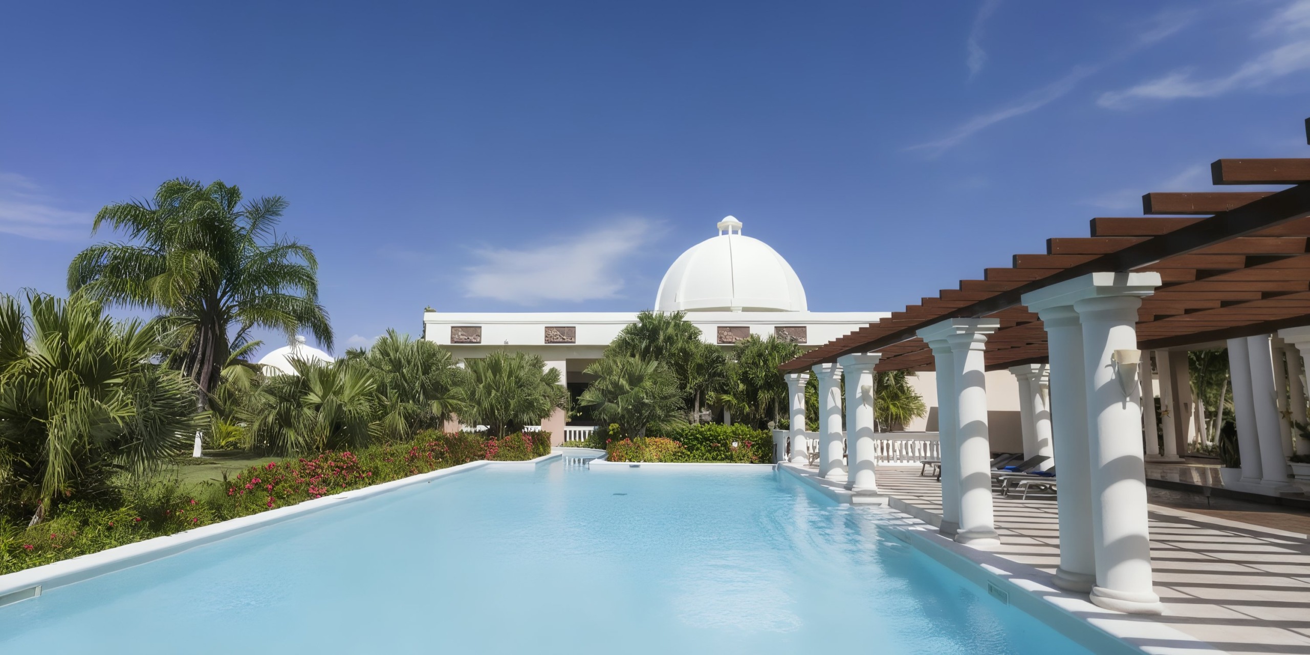 The crystal-clear outdoor swimming pool is surrounded by vibrant greenery, while the sophisticated white domed structure of the Grand Palladium Lady Hamilton provides a stunning backdrop beneath a bright blue sky.