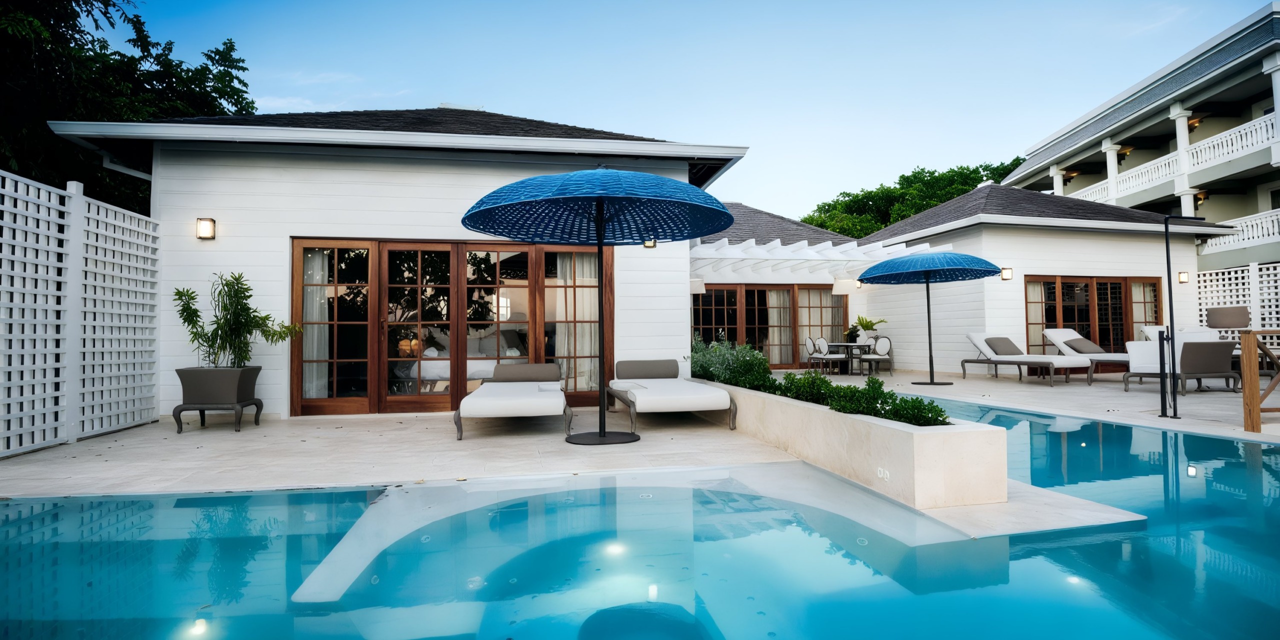 A modern villa featuring a pool, lounge chairs, and blue umbrellas mirrors the elegance of a Grand Palladium Lady Hamilton event. Its white exterior and glass doors exude sophistication, while a trellis frames the lush greenery against the backdrop of a clear blue sky.
