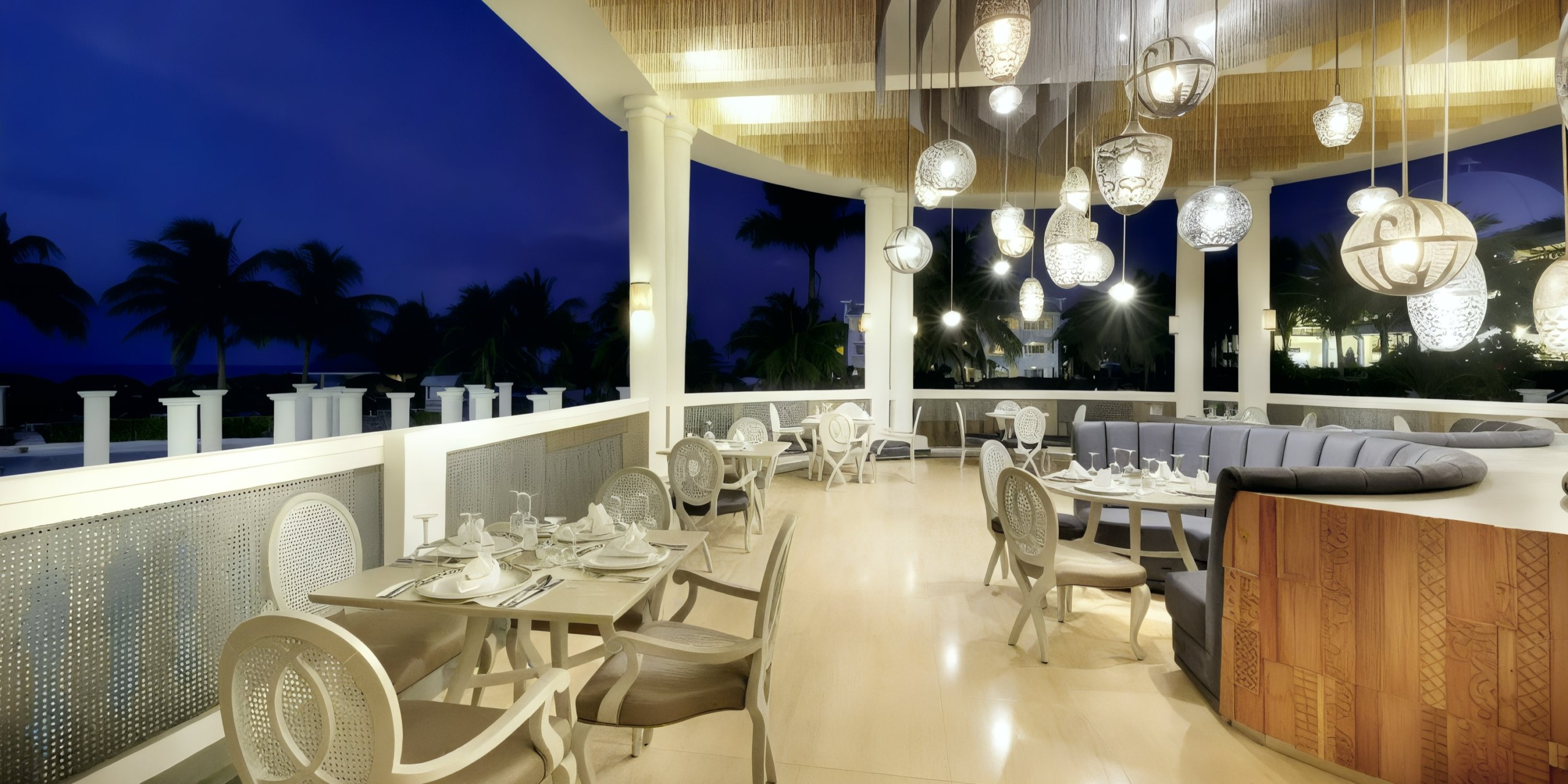 The Grand Palladium Lady Hamilton offers an elegant outdoor dining experience with round tables and cushioned seating, beautifully illuminated by hanging lanterns. This enchanting venue provides stunning views of palm trees under a deep blue evening sky, making it an ideal setting for a romantic wedding celebration.