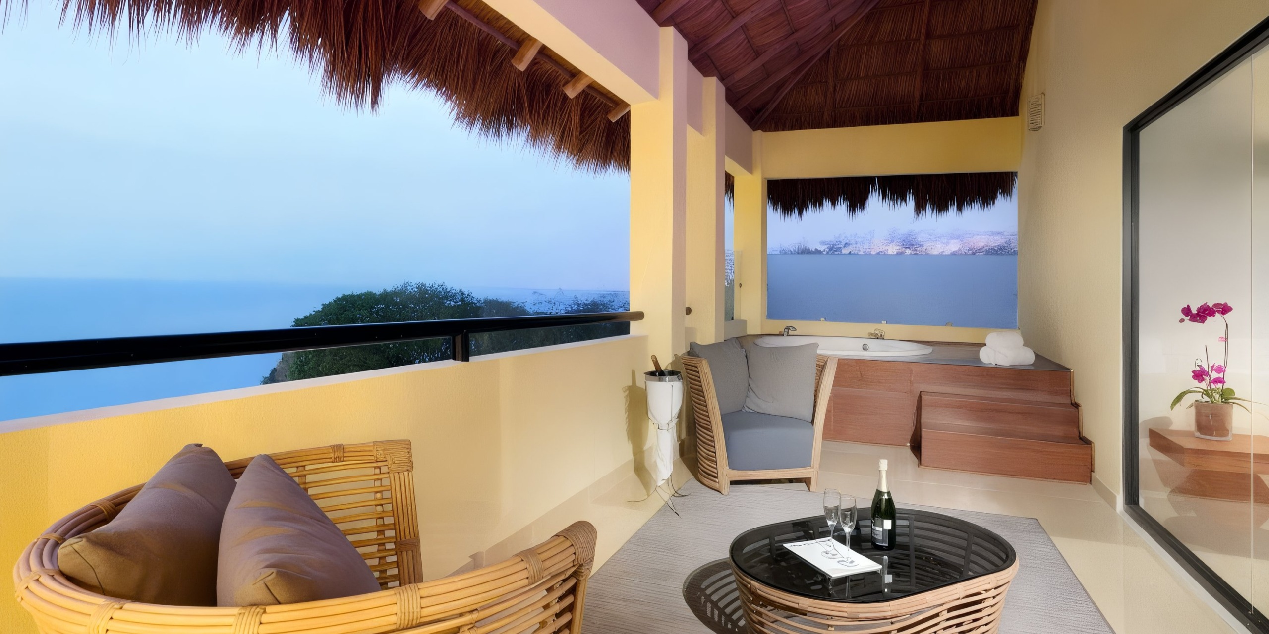 The balcony at the Grand Palladium Vallarta boasts wicker furniture, a small table complete with a bottle and glasses, an open book, a wooden hot tub, and a stunning view of the ocean.