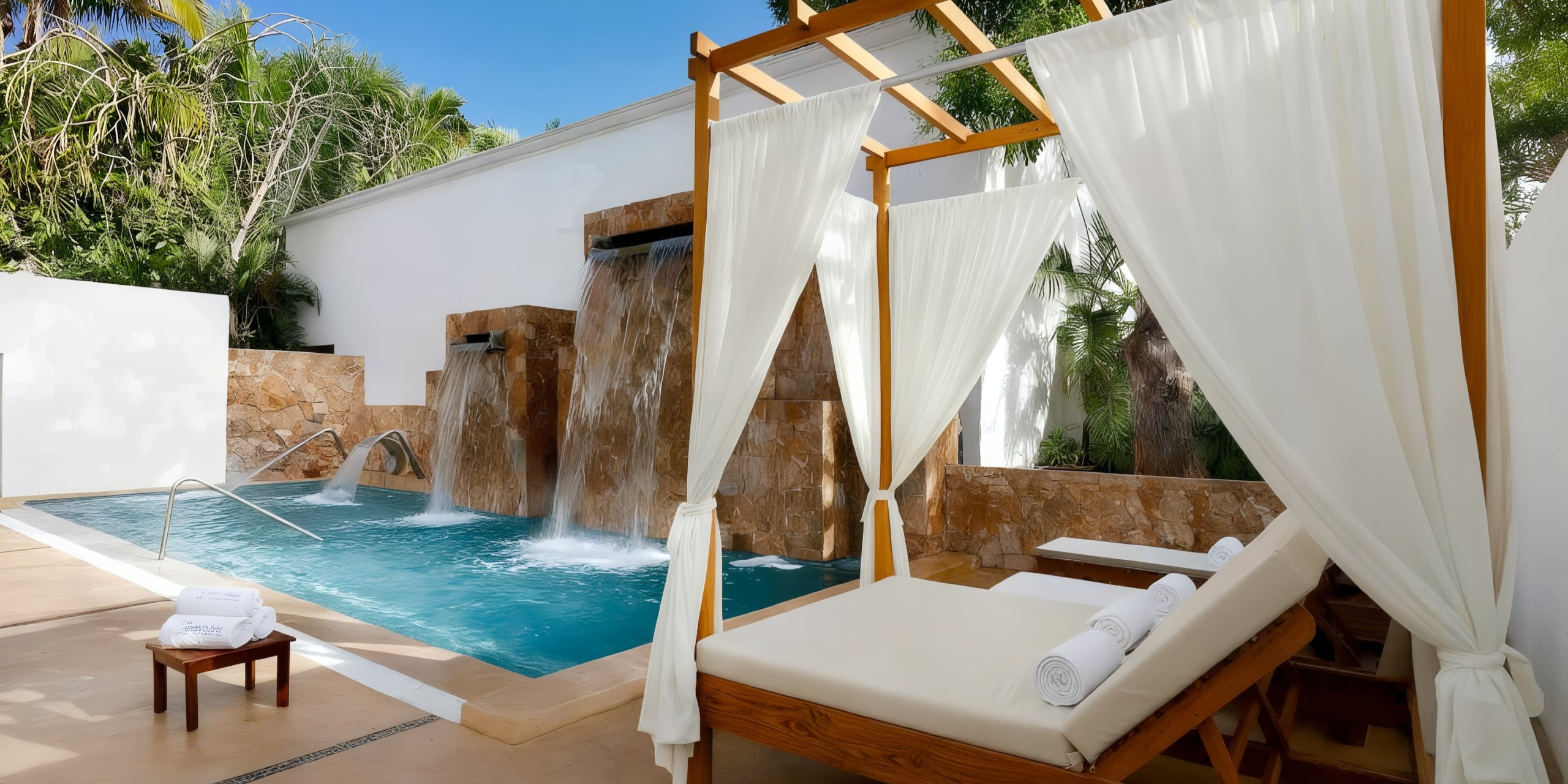 Experience the luxury of the Grand Palladium Vallarta's outdoor spa area, featuring a pool with water elements, a wooden lounge chair adorned with white fabric drapes, and neatly rolled towels. All of this is beautifully surrounded by lush greenery and elegant stone accents.