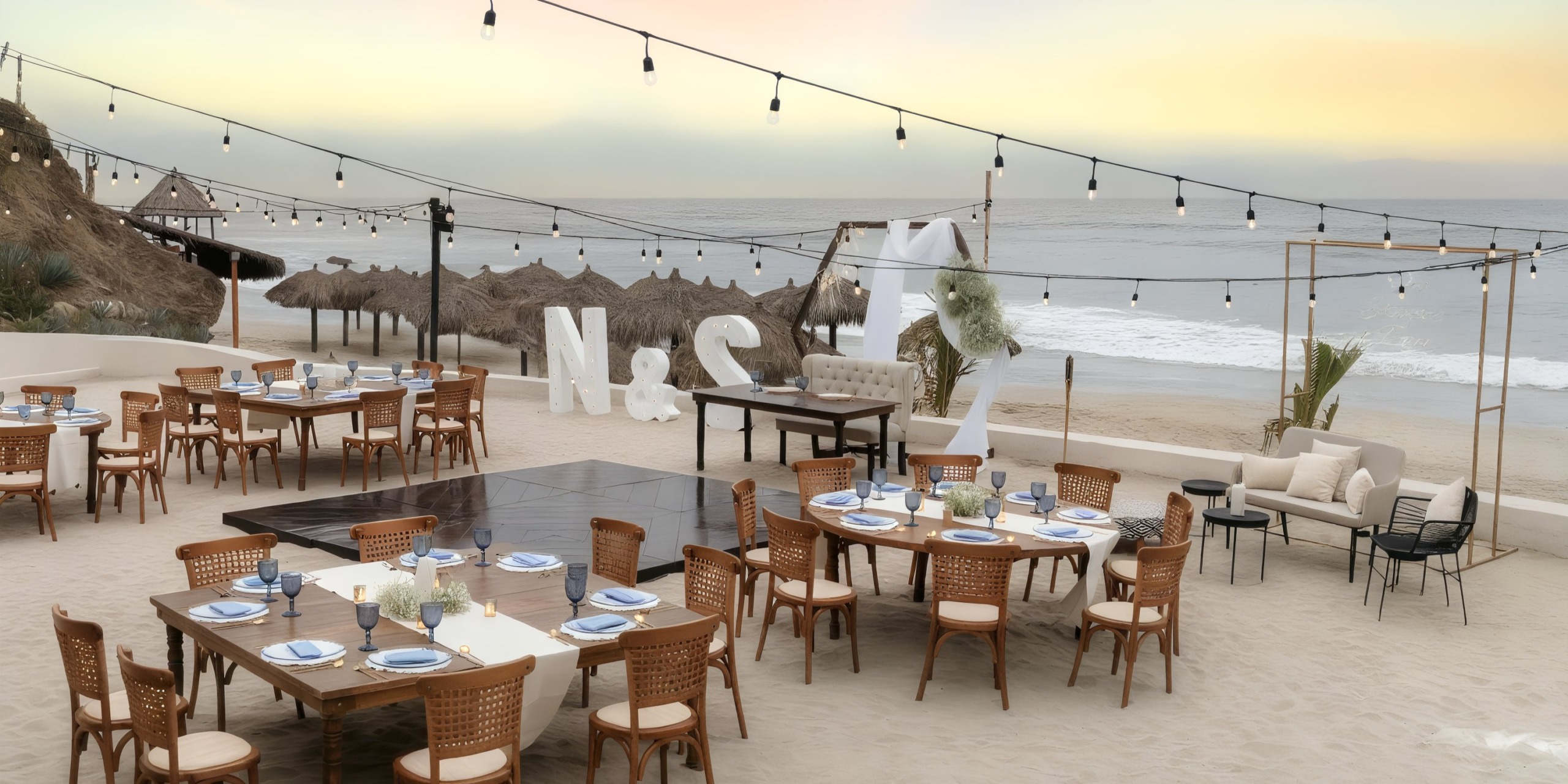 Beachside dining setup at the Grand Palladium Vallarta, featuring wooden tables and chairs adorned with string lights. Large letters "N" and "S" are positioned near the ocean.