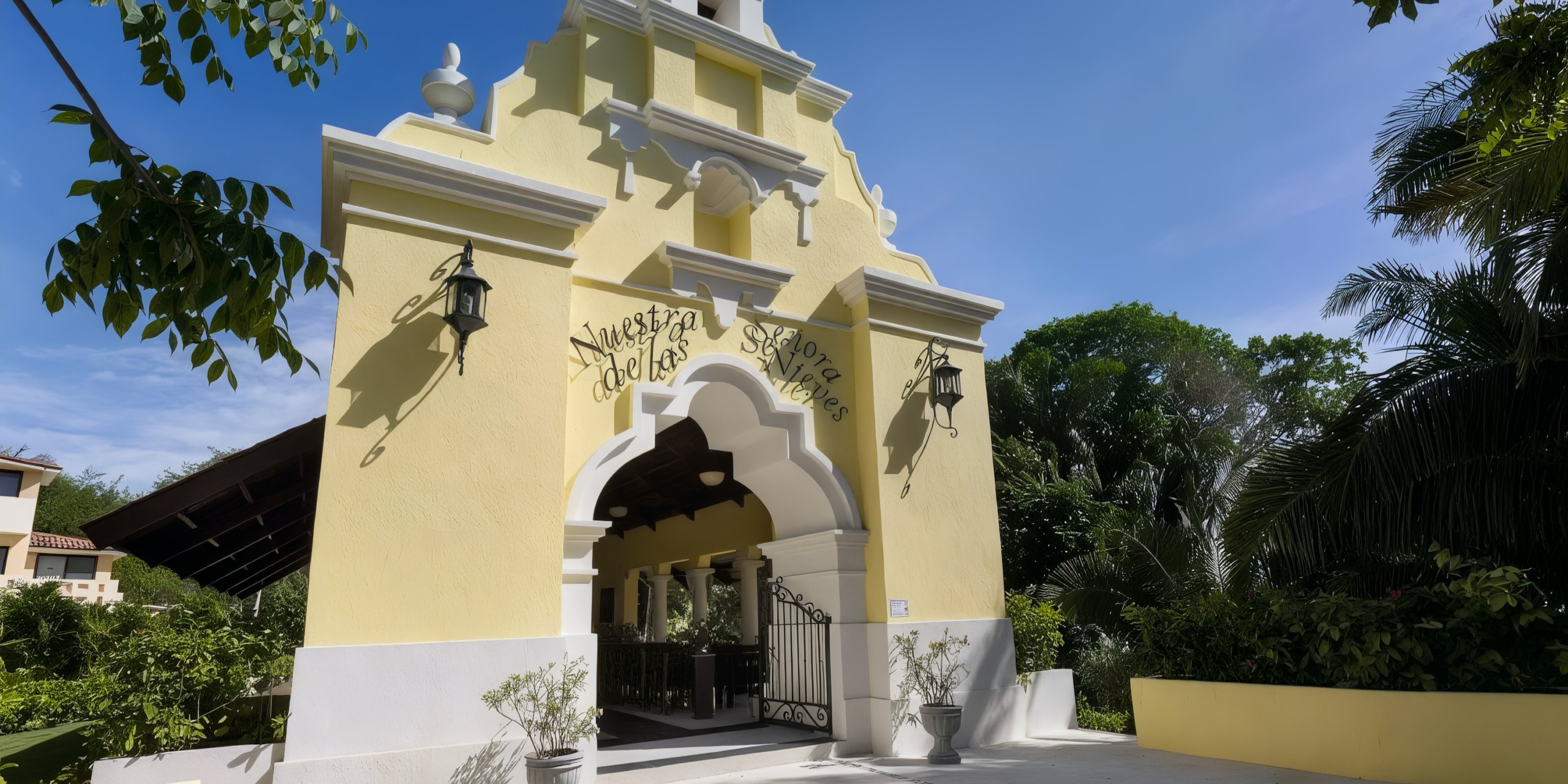 The Grand Palladium Vallarta, with its vibrant yellow facade and ornate entrance, is beautifully surrounded by lush greenery under a clear blue sky.