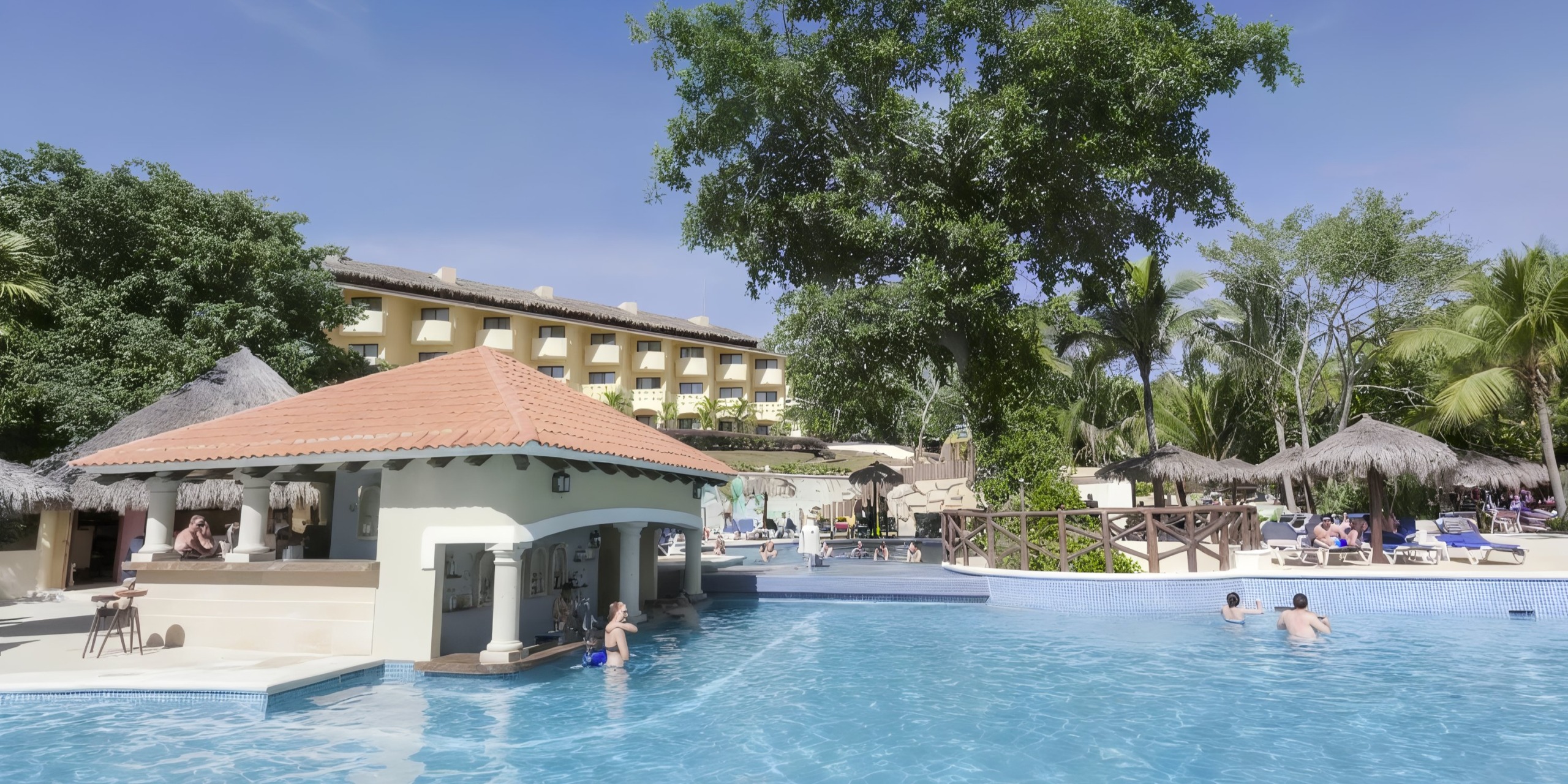 At the Grand Palladium Vallarta, enjoy a resort pool scene complete with a swim-up bar, guests relaxing in the water, and a hotel building framed by lush tropical trees in the background.