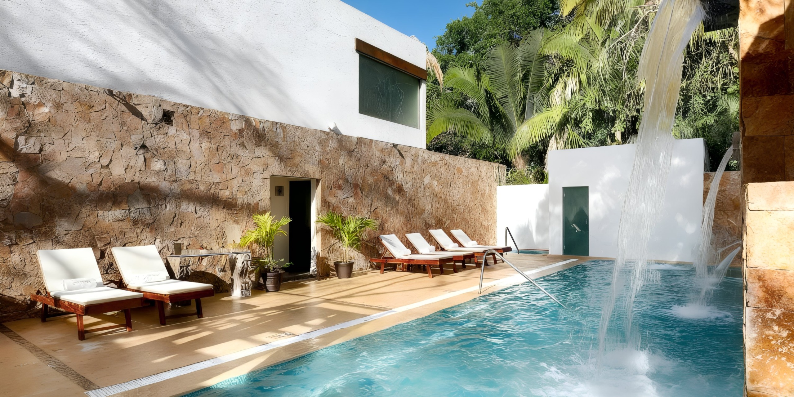 Enjoy the poolside area at Grand Palladium Vallarta, featuring lounge chairs, a stone wall, and lush tropical plants. A stunning waterfall feature flows into the pool for a serene experience.