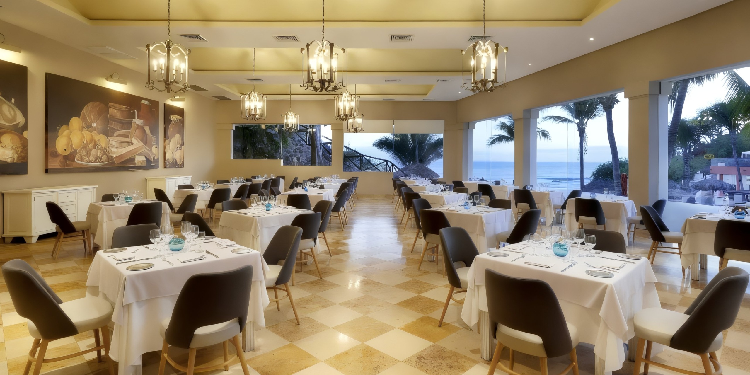 The Grand Palladium Vallarta features an elegant setting with neatly arranged tables and chairs, chandeliers, large paintings, and wide windows offering a stunning view of the ocean.