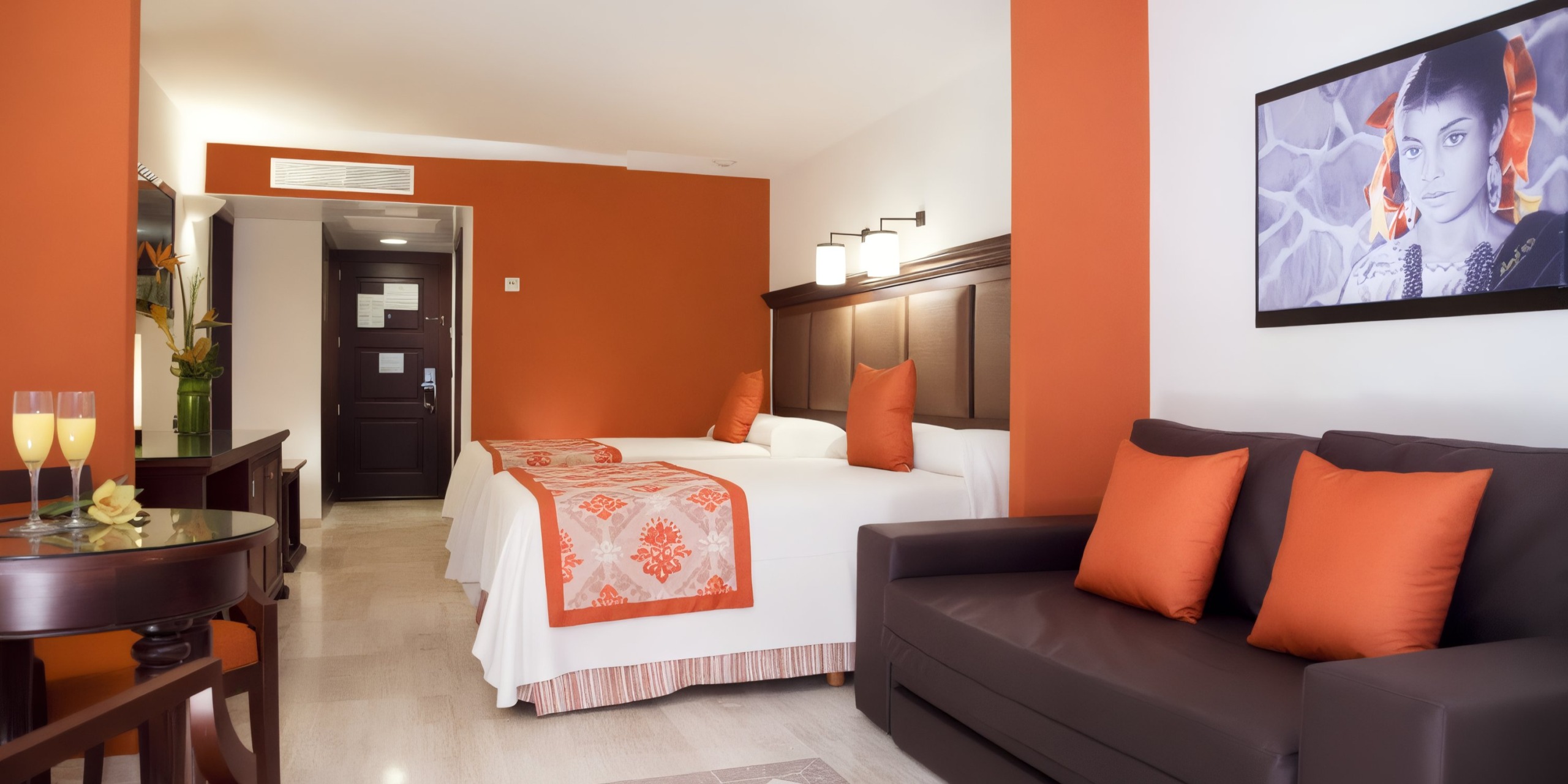 In a Grand Palladium Vallarta hotel room, you'll find two beds complemented by orange and white decor, a sofa adorned with orange cushions, a small table stocked with drinks, and a wall-mounted monochrome portrait.