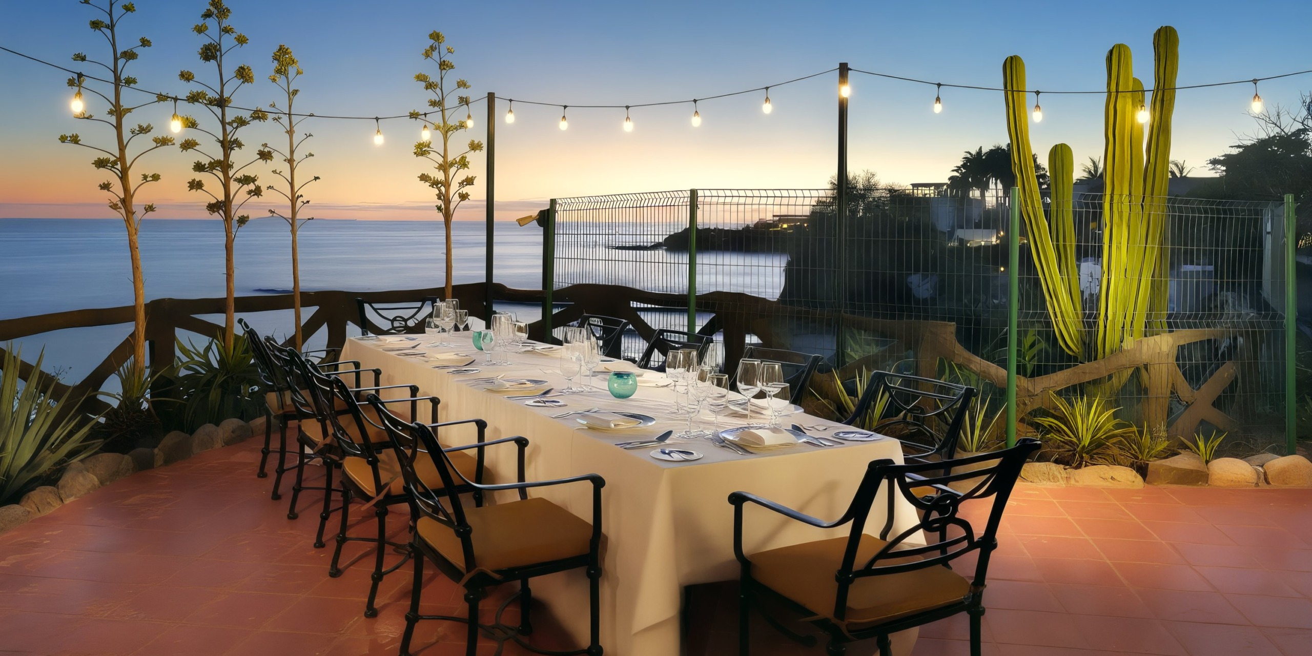 Enjoy a picturesque outdoor dining experience at the Grand Palladium Vallarta, featuring an elegant long table and chairs on a terrace with breathtaking ocean views at sunset. String lights are suspended overhead, and large cacti accentuate the surroundings.