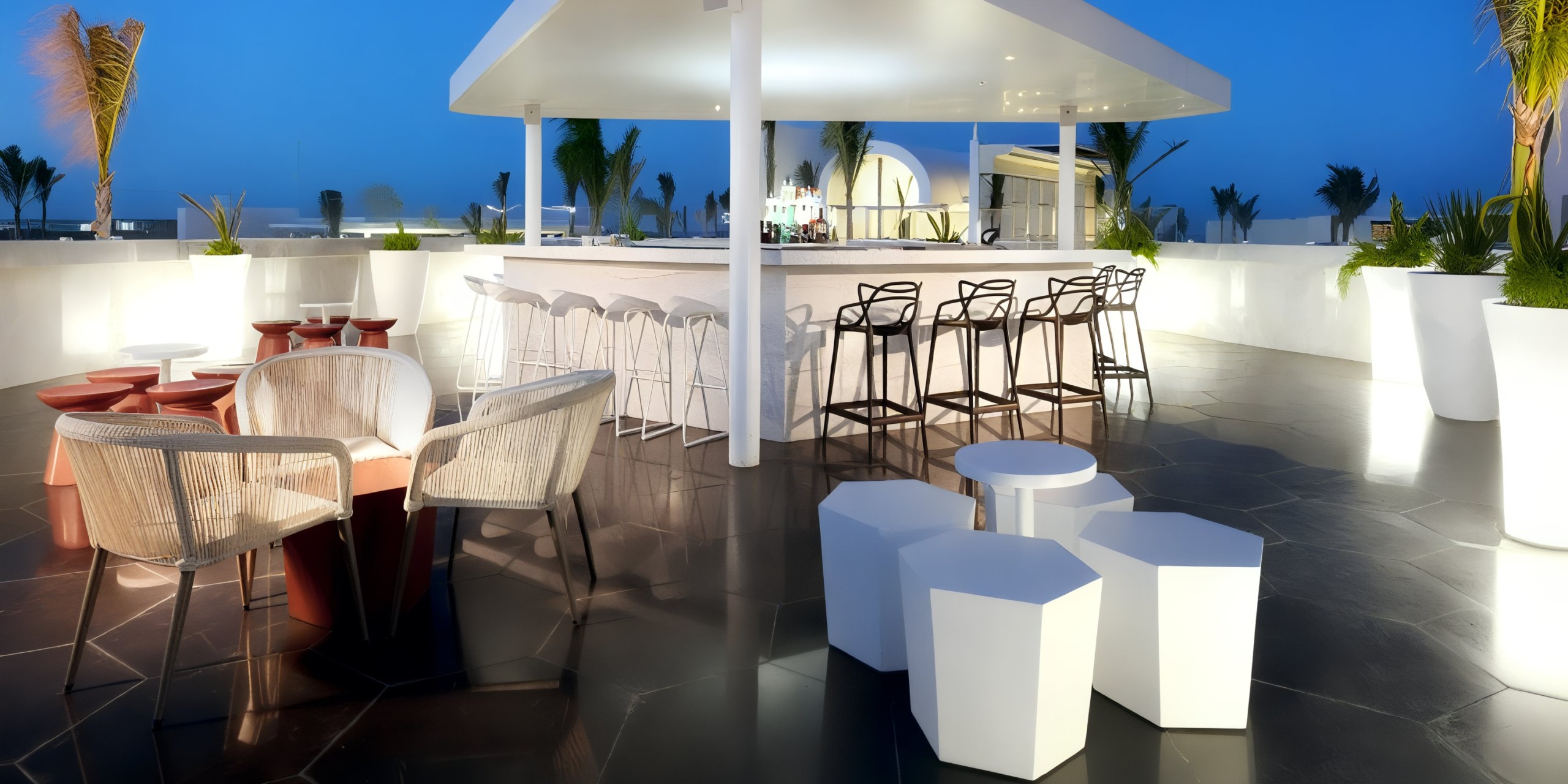 The Grand Palladium Costa Mujeres features a rooftop bar adorned with sleek white furniture, high chairs lining the counter, and lush potted plants, all set against a backdrop of deep blue sky.