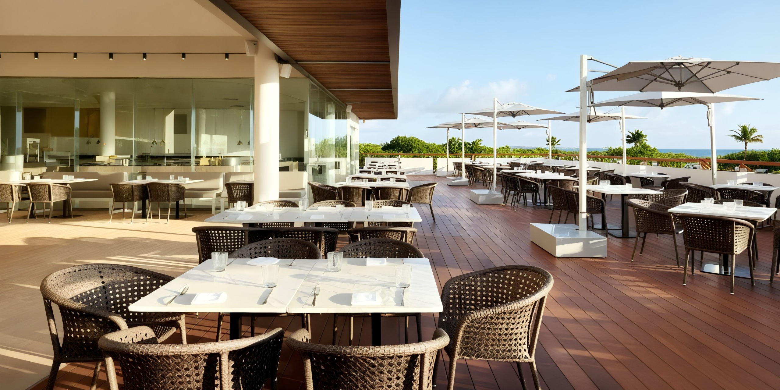 The Grand Palladium Costa Mujeres features an outdoor restaurant patio adorned with wicker chairs and white tables. The space is beautifully shaded by large umbrellas, while glass walls and wooden flooring enclose the area. A partly cloudy sky complements the lush greenery in the background.