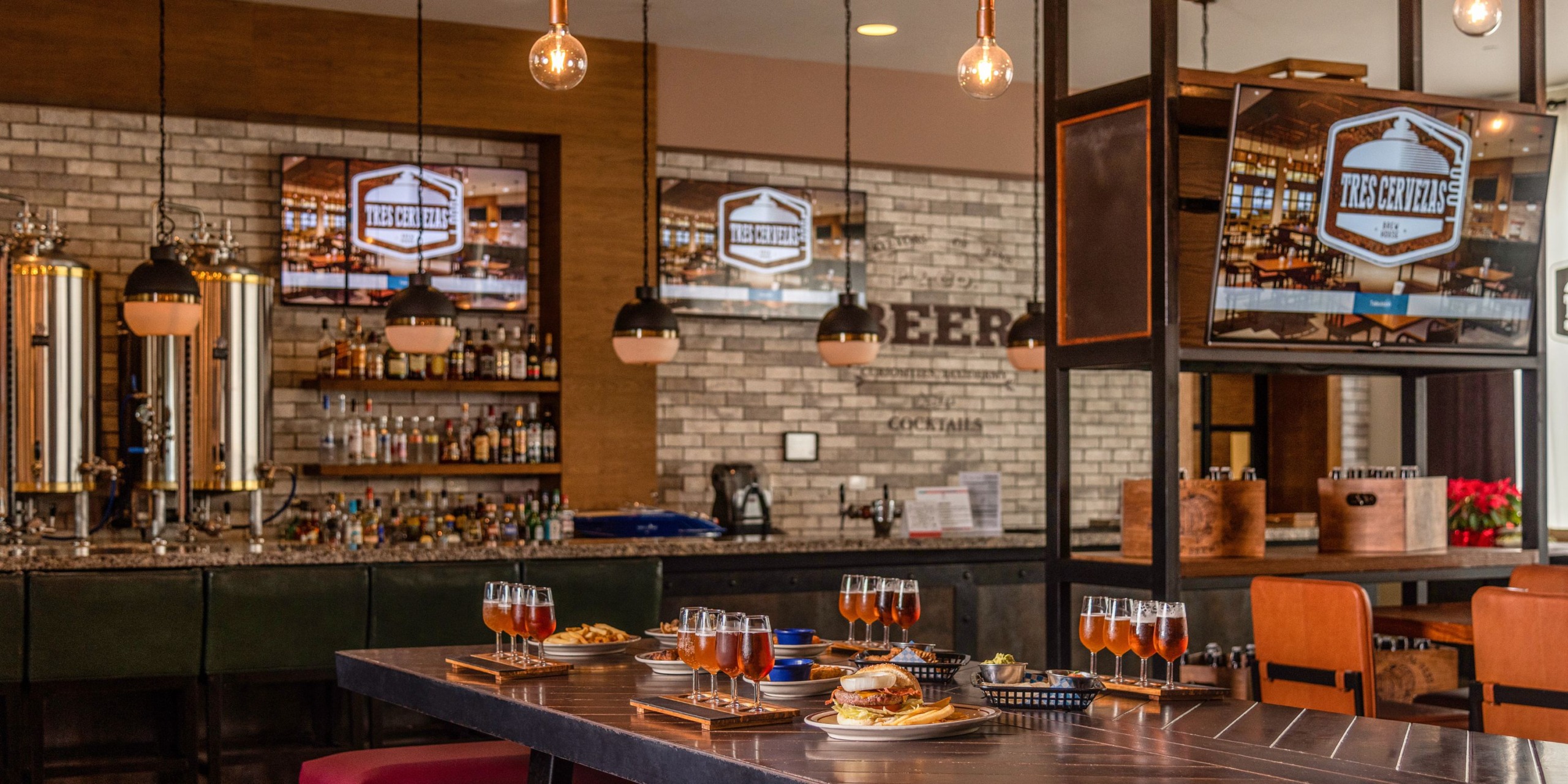 A contemporary setting at Hyatt Ziva Cancun features tables adorned with flights of beer and burgers, while TVs showcase the Hyatt Ziva Cancun logo.