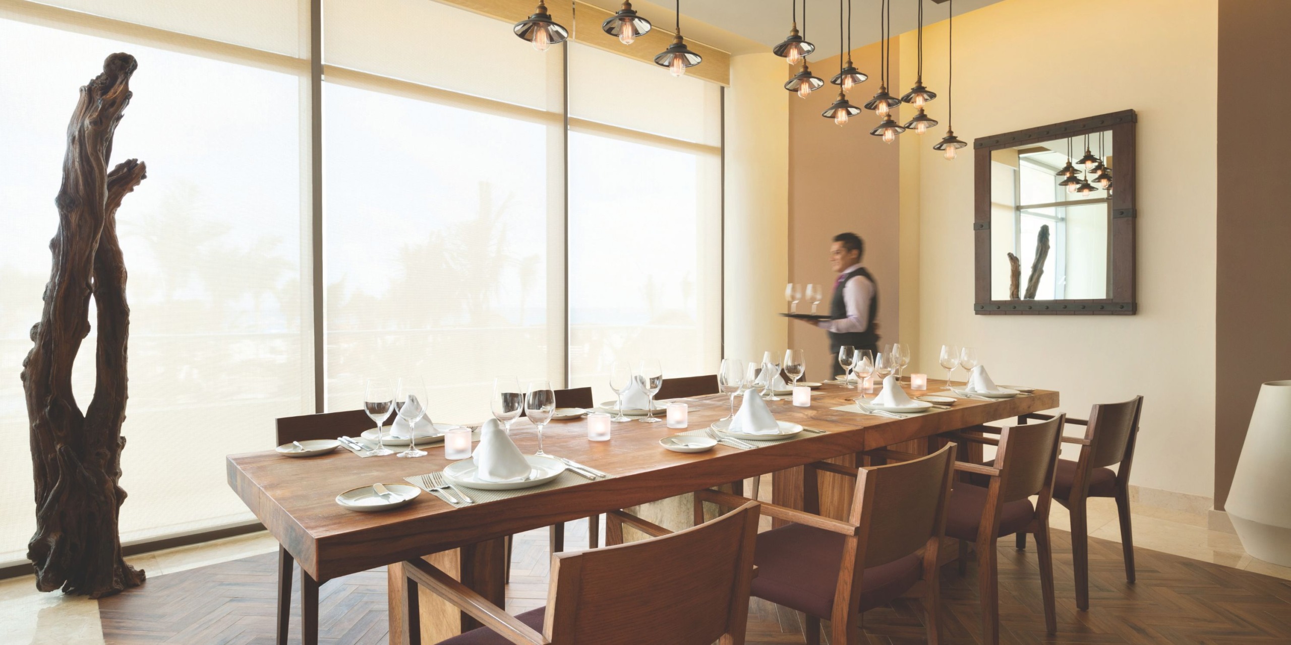 The Hyatt Ziva Cancun dining room is elegantly furnished with a wooden table set for eight, featuring neatly arranged plates, glasses, and folded napkins. In the background, a waiter carries additional glasses. The space is adorned with a large mirror and an exquisite sculpture.